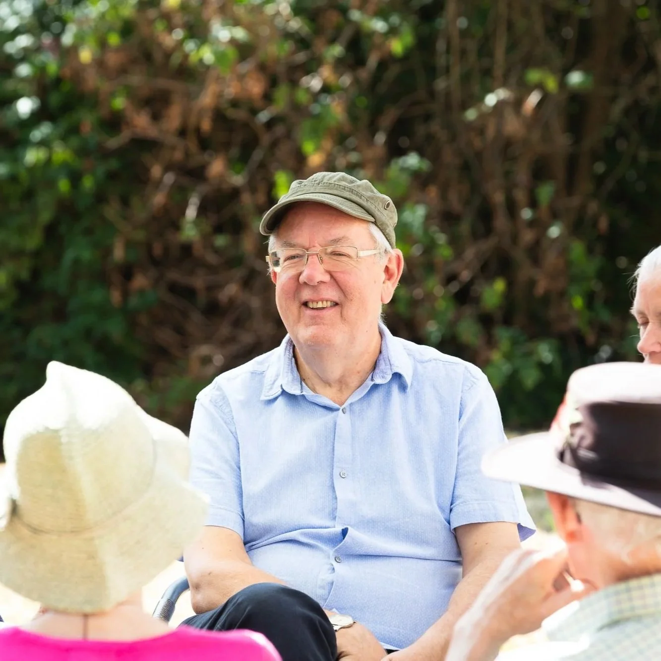 A group of elderly people sitting outdoors, with one man smiling and wearing a beige cap and light blue shirt, surrounded by others wearing hats.