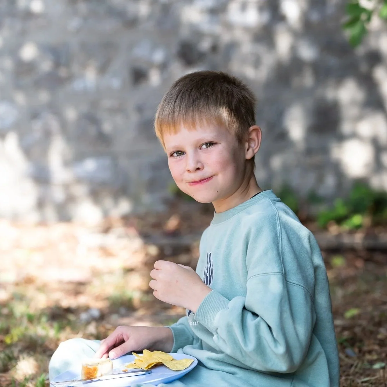 A young boy with short brown hair and fair skin sitting outdoors, smiling at the camera, holding a plate of food with potato chips and a sandwich, with a blurred background of trees and a stone or concrete wall.