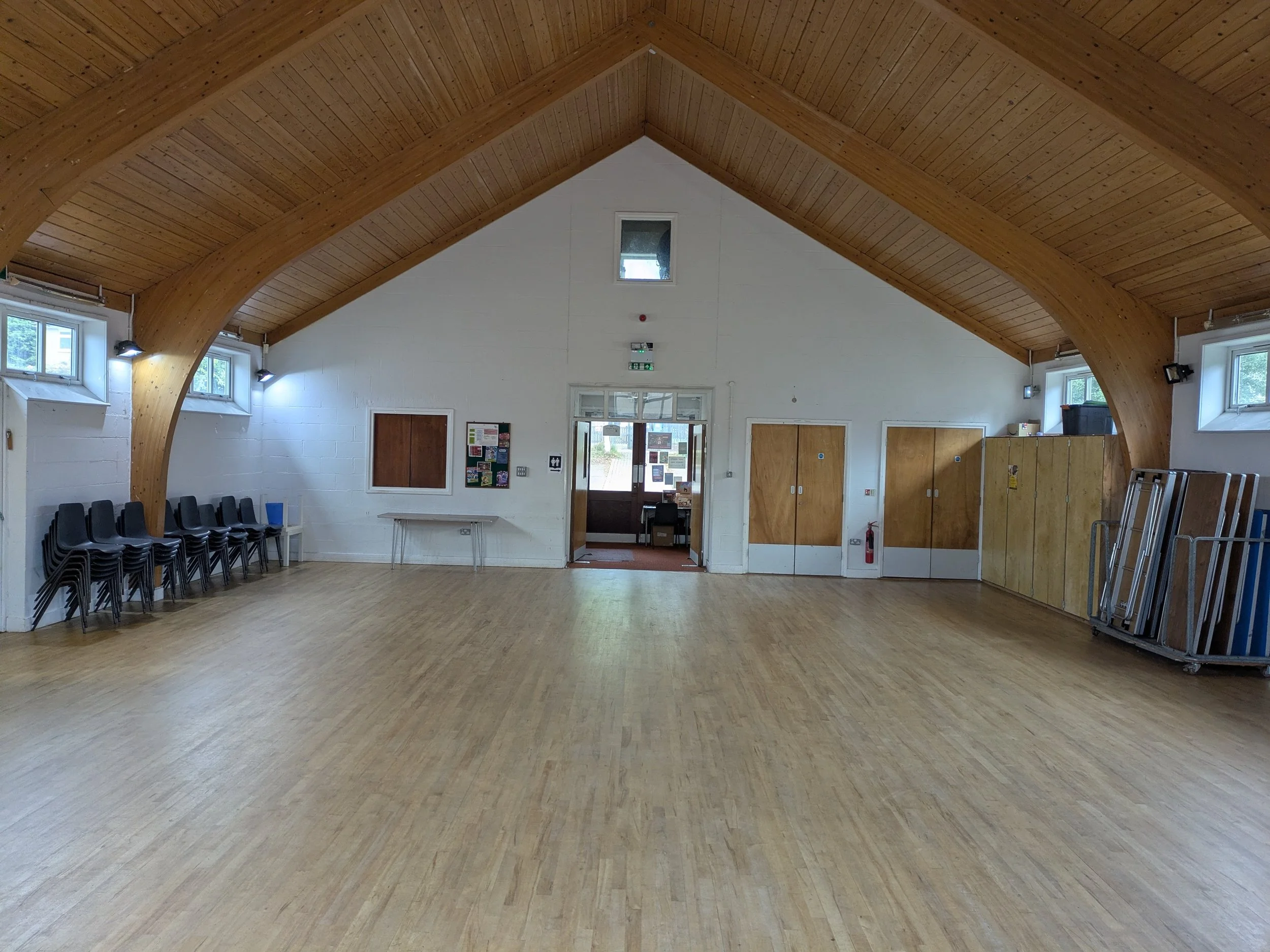 An interior view of a large, empty community hall or event space with a high, wooden vaulted ceiling, white walls, multiple small windows, and a wood floor. There are chairs lined up along one wall and some equipment and furniture on the right side.