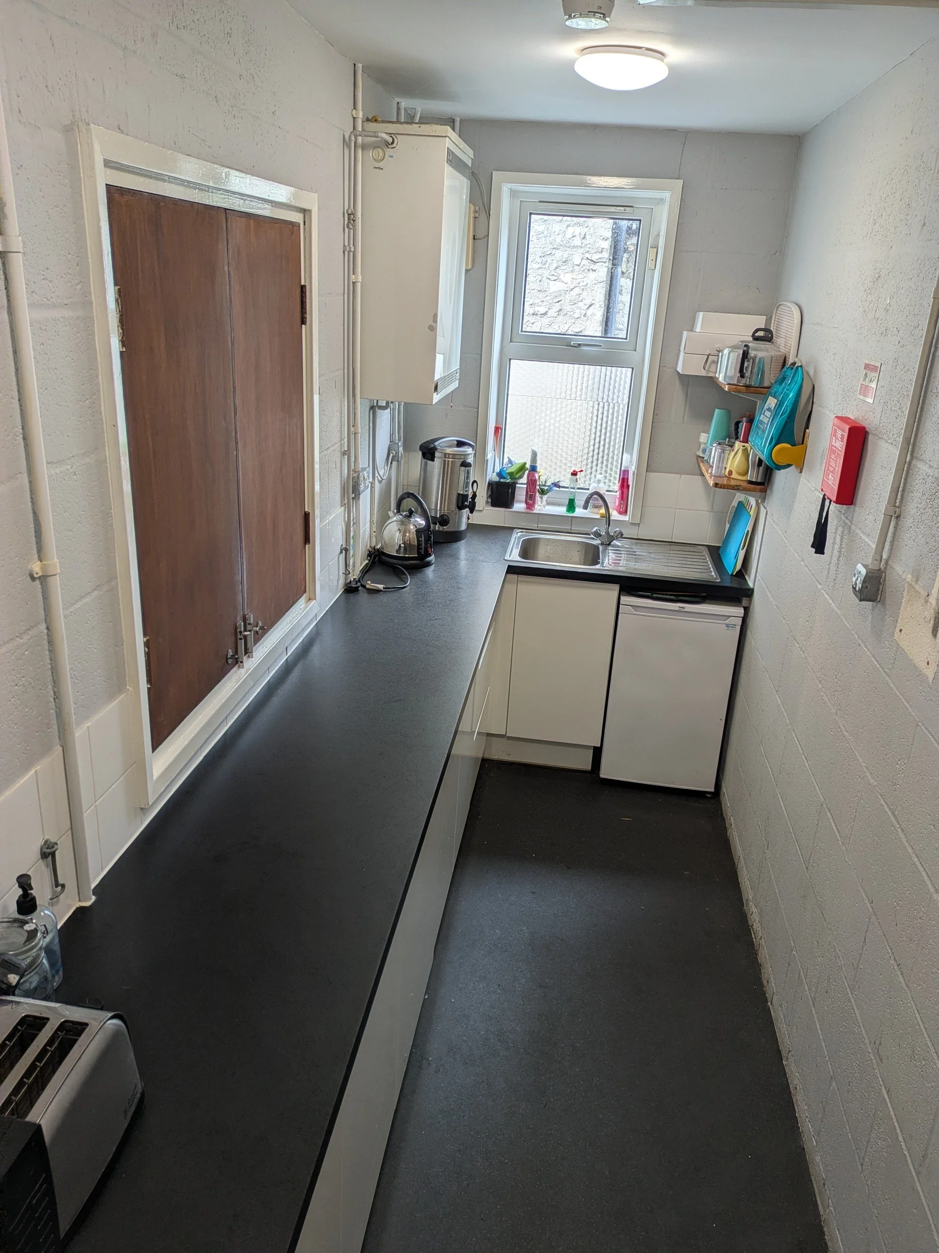 A small kitchen with a black countertop, a sink with a window above it showing a stone wall outside, shelves with dishes and utensils, a toaster oven, a kettle, an electric water boiler, and various cleaning supplies, with white walls and a ceiling l