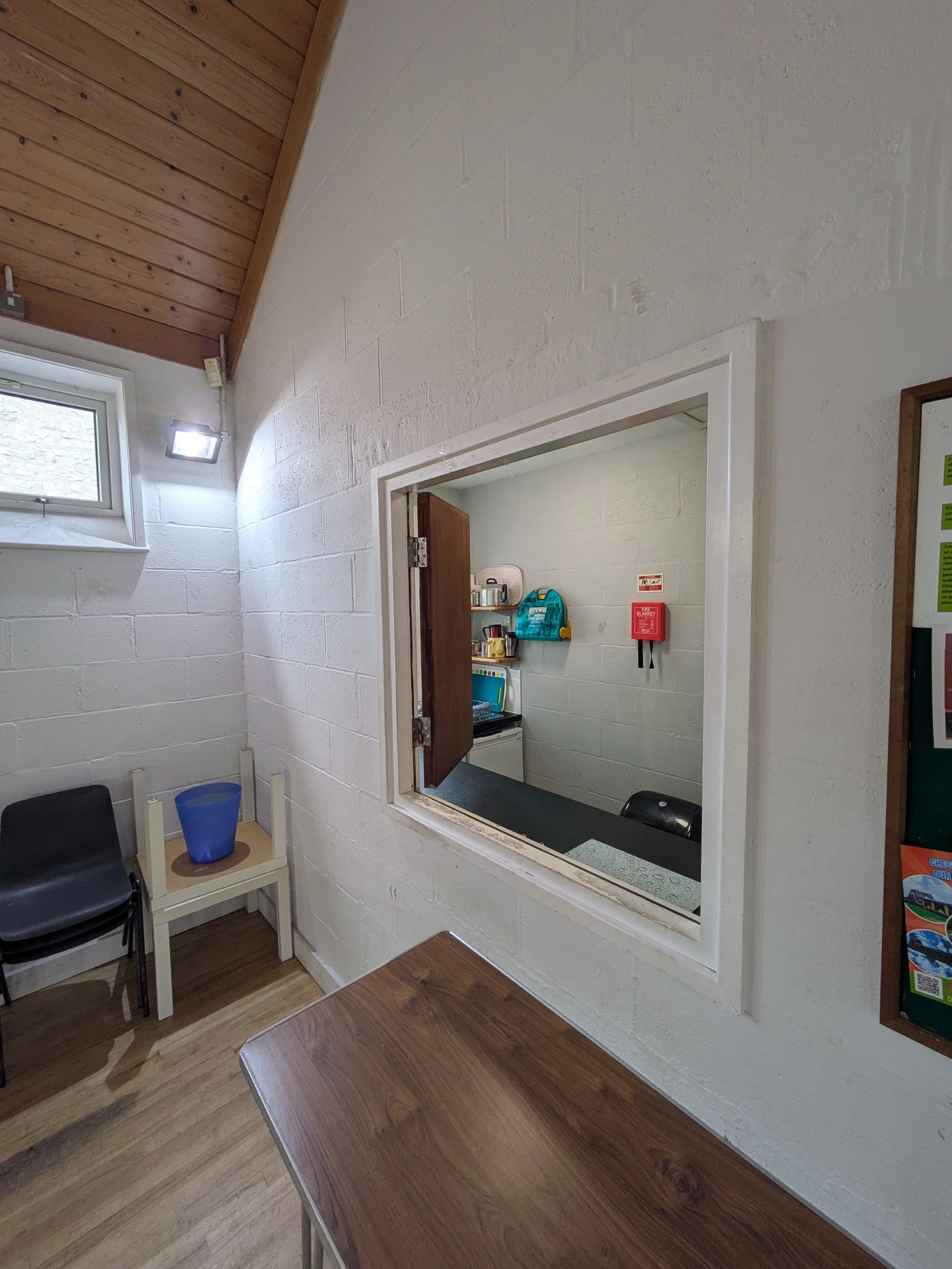 Interior of a room with white brick walls, a small window, and wooden ceiling. There is a table with a wooden top and a black chair, and a small window opening to an office or reception area with shelves and various items.