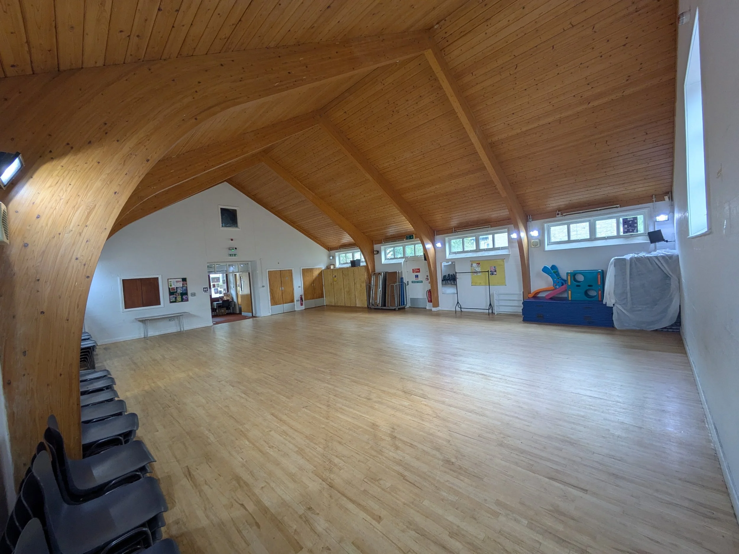 Interior view of a spacious room with a high, curved wooden ceiling, multiple windows along the walls, a stack of blue and orange children's play equipment, chairs, and a white covered object.