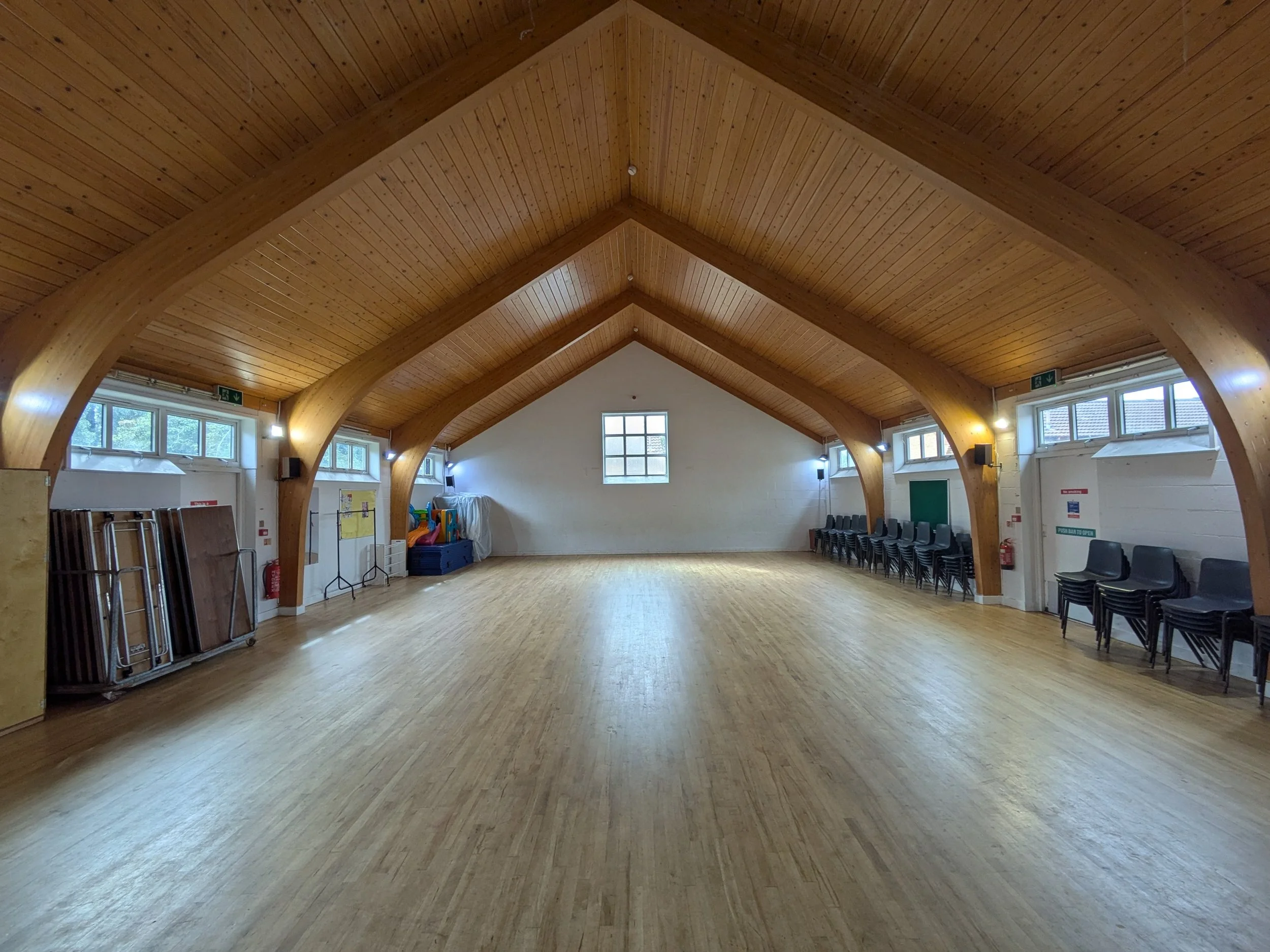 A spacious community hall with a high wooden vaulted ceiling, large windows along the walls, and a wooden floor. There are stacked chairs and some stored equipment along the sides of the room.