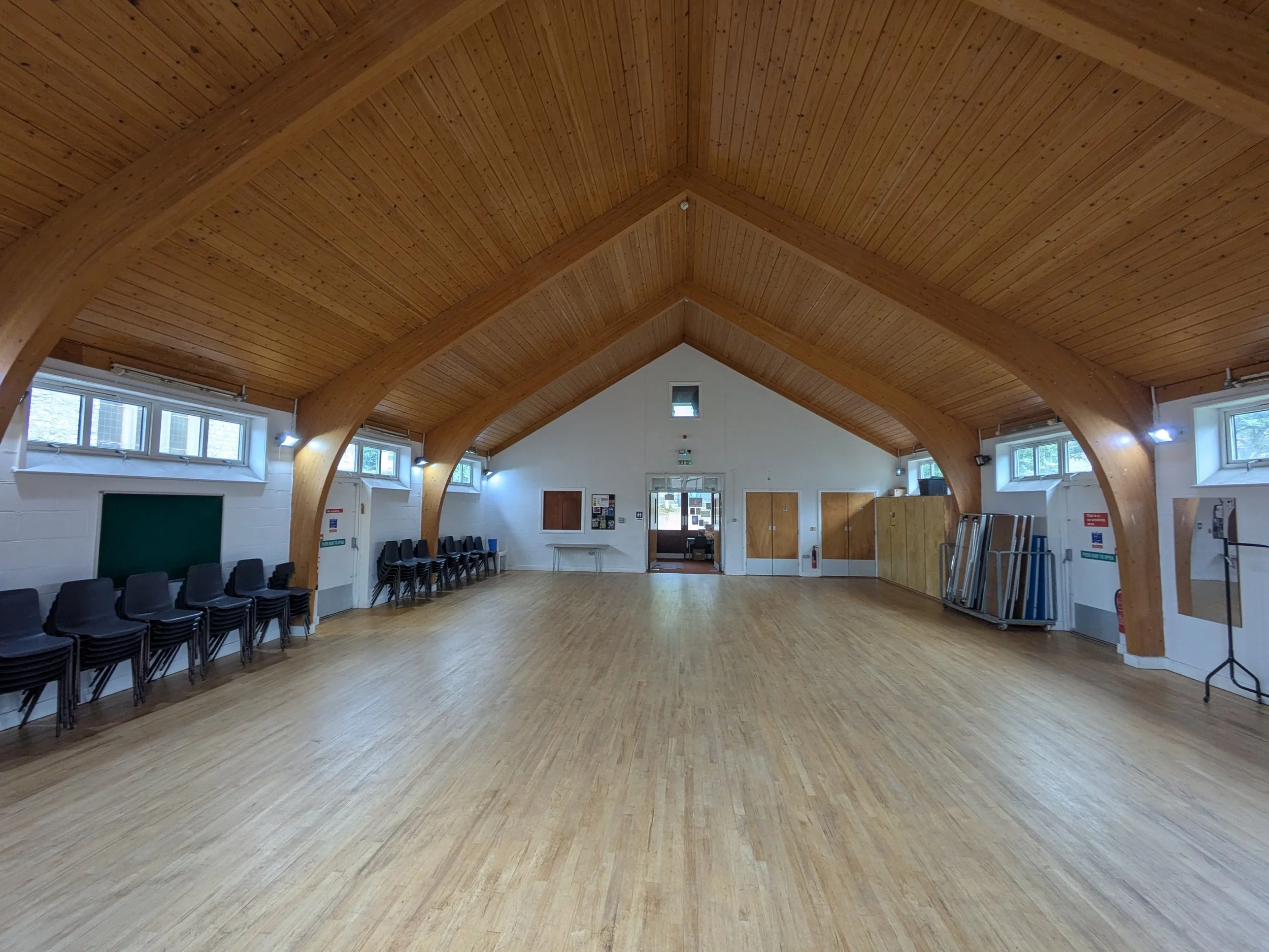An indoor hall with a high wood-paneled ceiling, several windows, chairs along the walls, and a wooden floor.