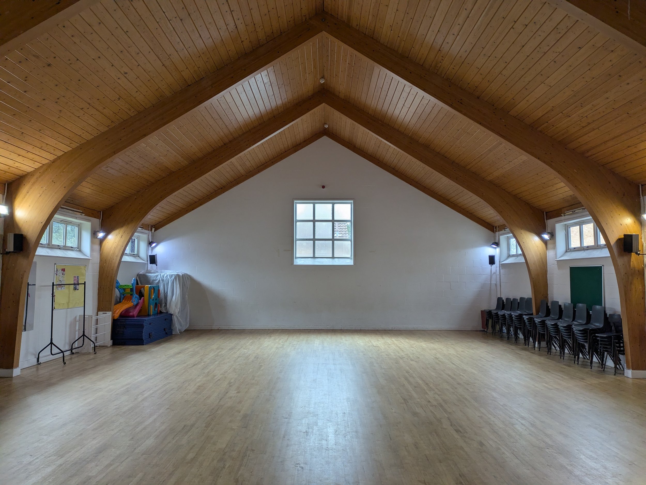 Empty community hall with wooden arched ceiling, large window, chairs stacked on the side, and some musical equipment.