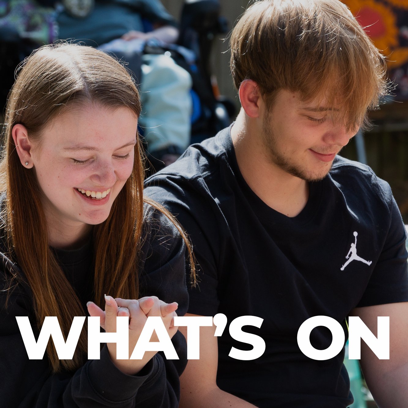 A young woman and a young man sitting outdoors, smiling and looking at something. The woman has long red hair, freckles, and is wearing a black hoodie. The man has light brown hair, a beard, and is wearing a black T-shirt with a white Jumpman logo.