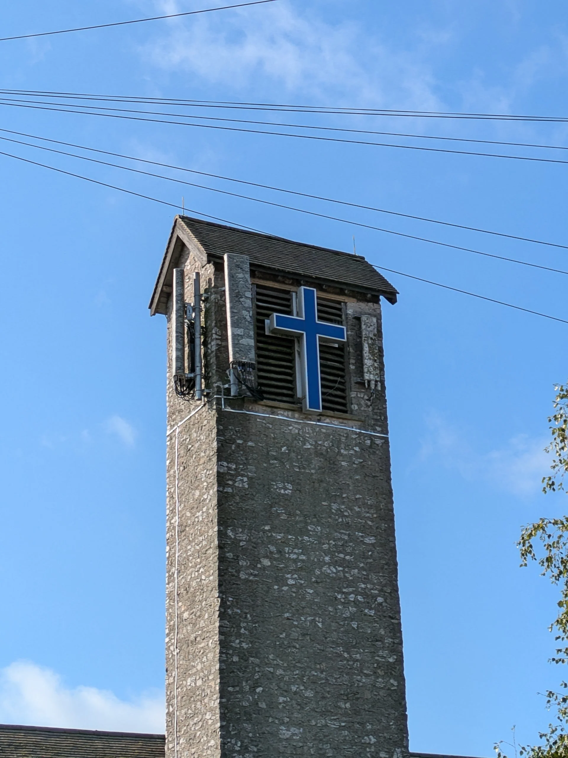 A church bell tower made of stone with a blue cross sign. The tower is topped with a small roof under a blue sky with a few clouds. Overhead power lines are visible across the sky.