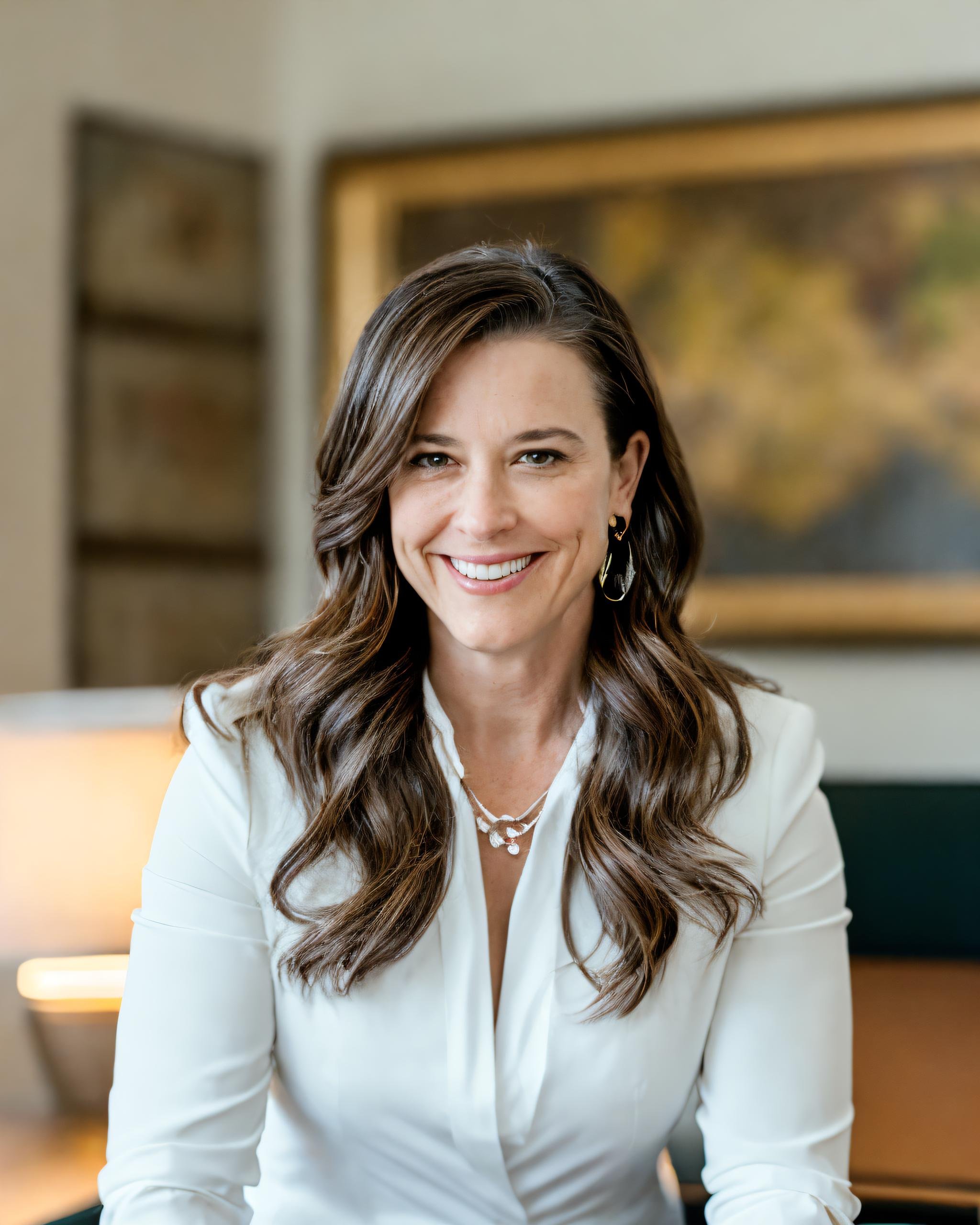 A woman with long brown wavy hair and earrings smiling at the camera, wearing a white blouse, in a room with artwork and a window in the background.