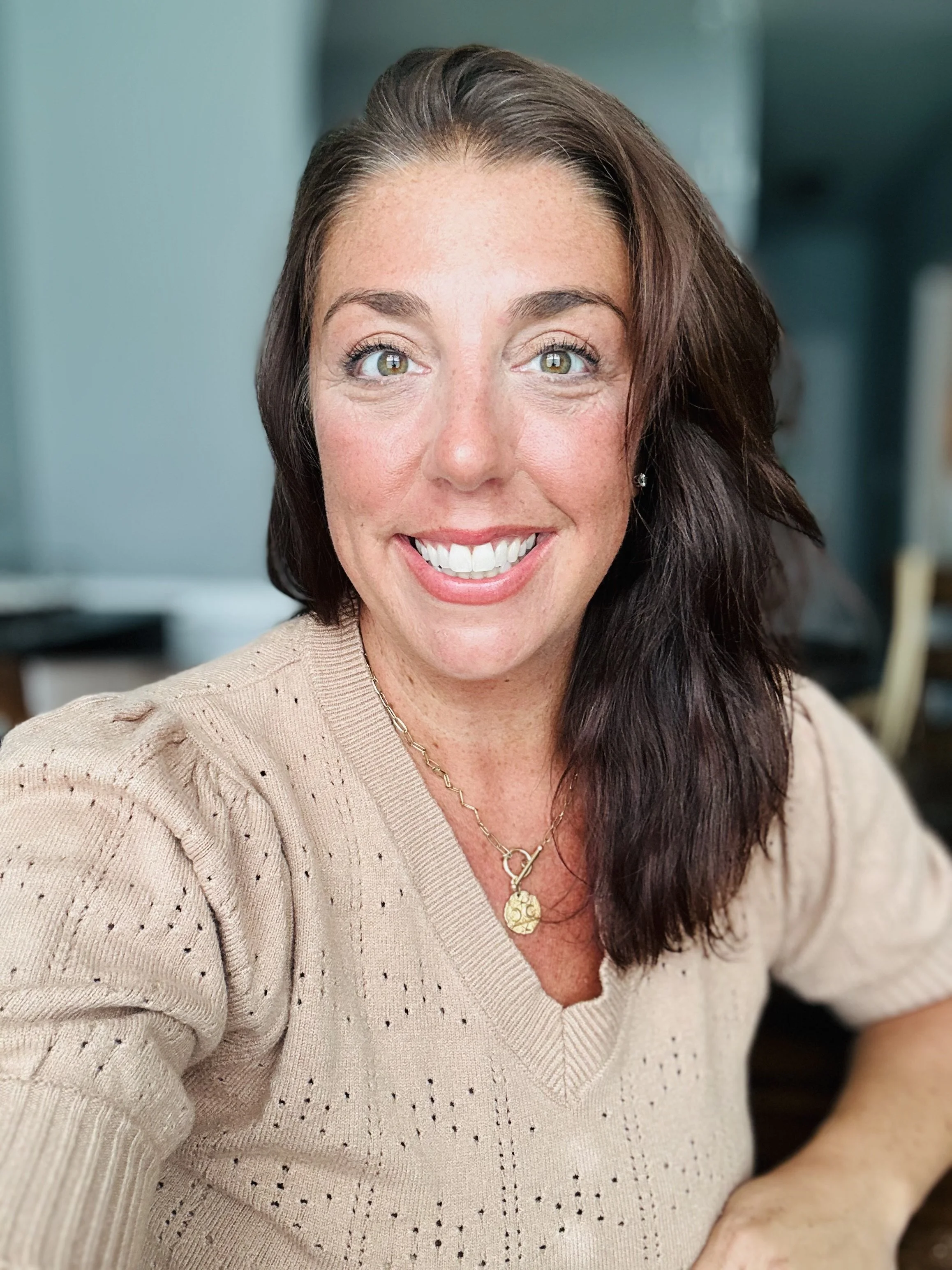 Close-up photograph of a woman with dark brown hair, smiling, wearing a beige sweater and gold jewelry, in an indoor setting with a blurred background.