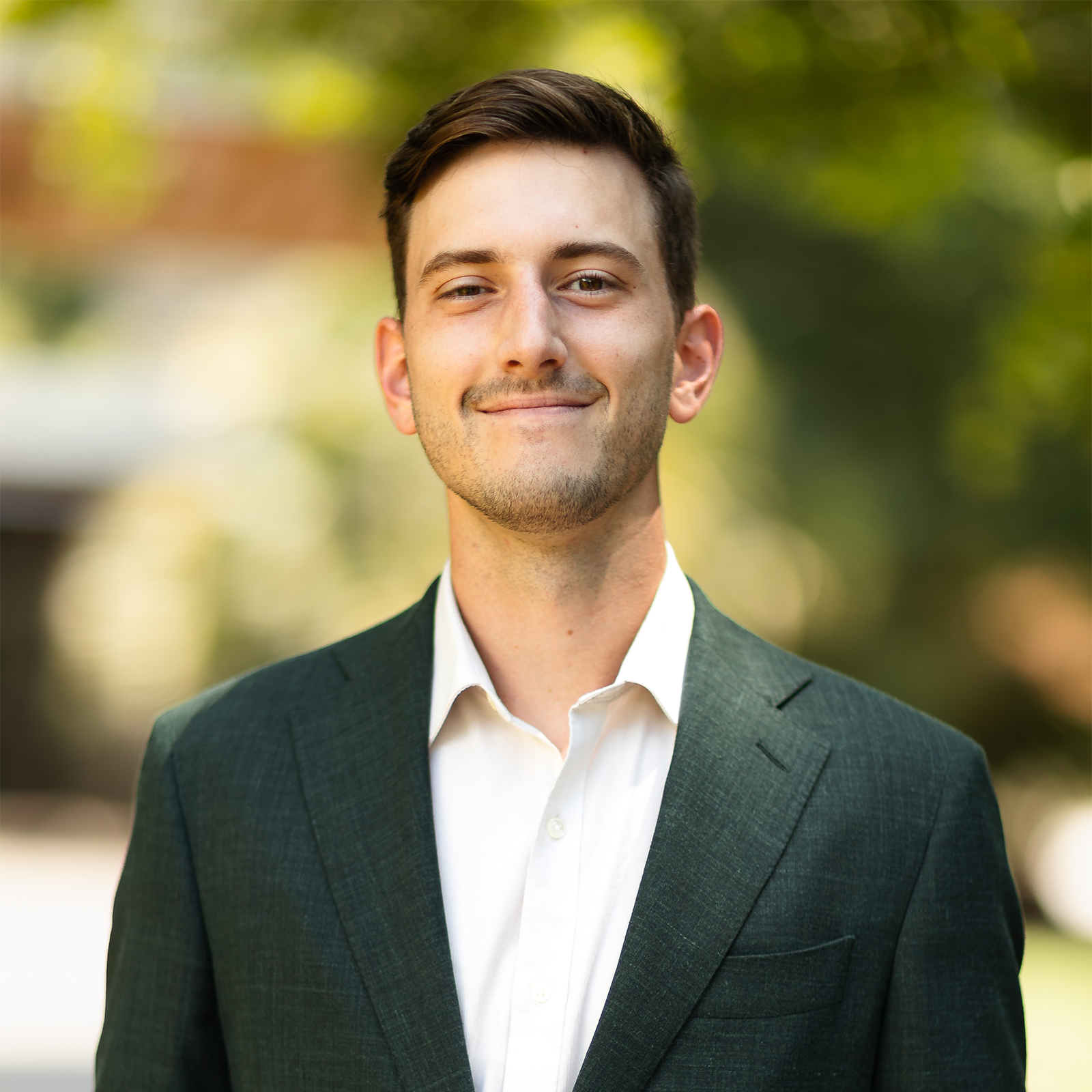 A young man with short brown hair, smiling, dressed in a white shirt and dark grey suit, standing outdoors with blurred green trees in the background.
