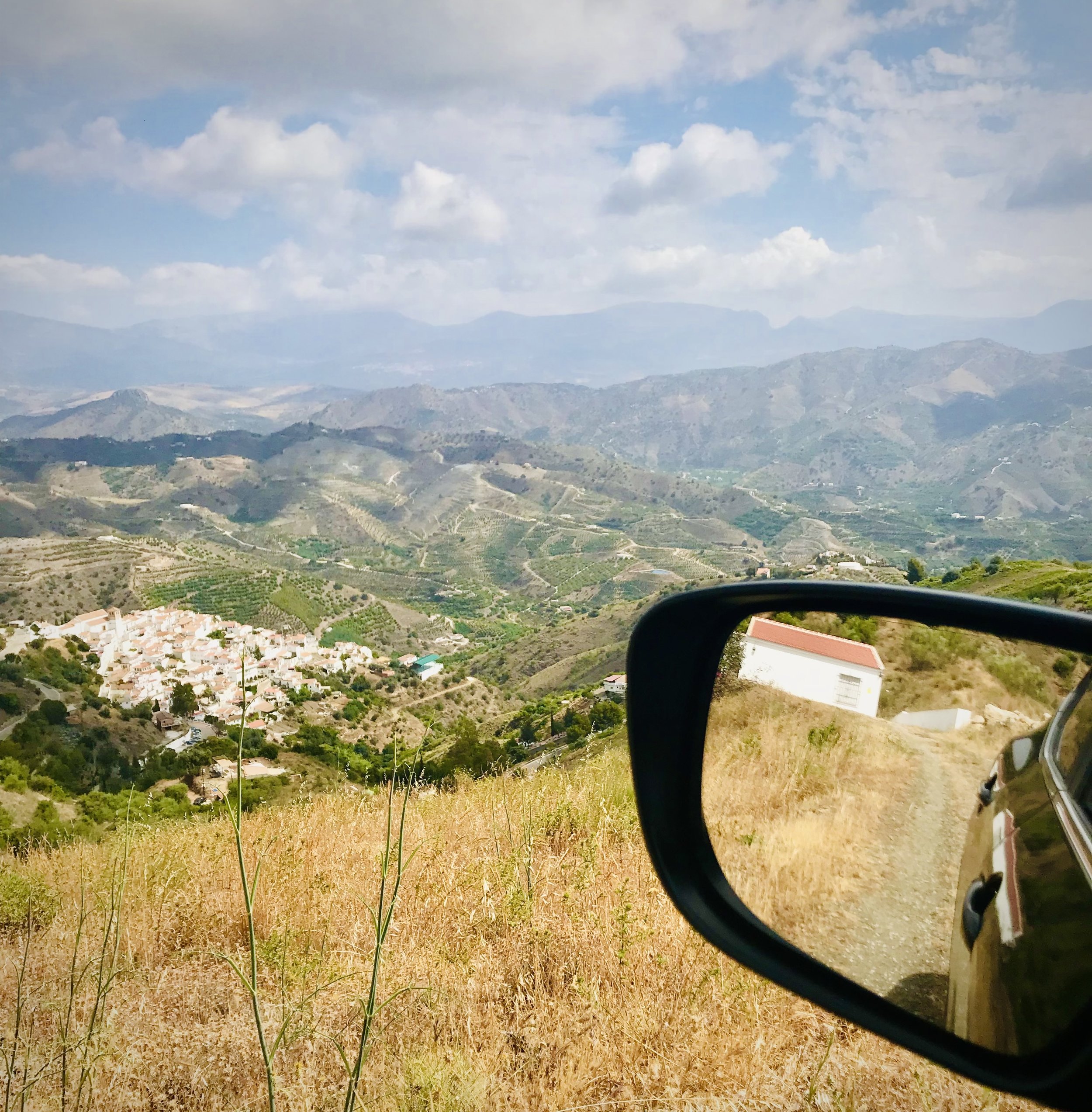 A scenic view of mountains in Andalucia with partly cloudy skies above and a small town in the foreground, viewed from a vehicle with a side mirror visible.