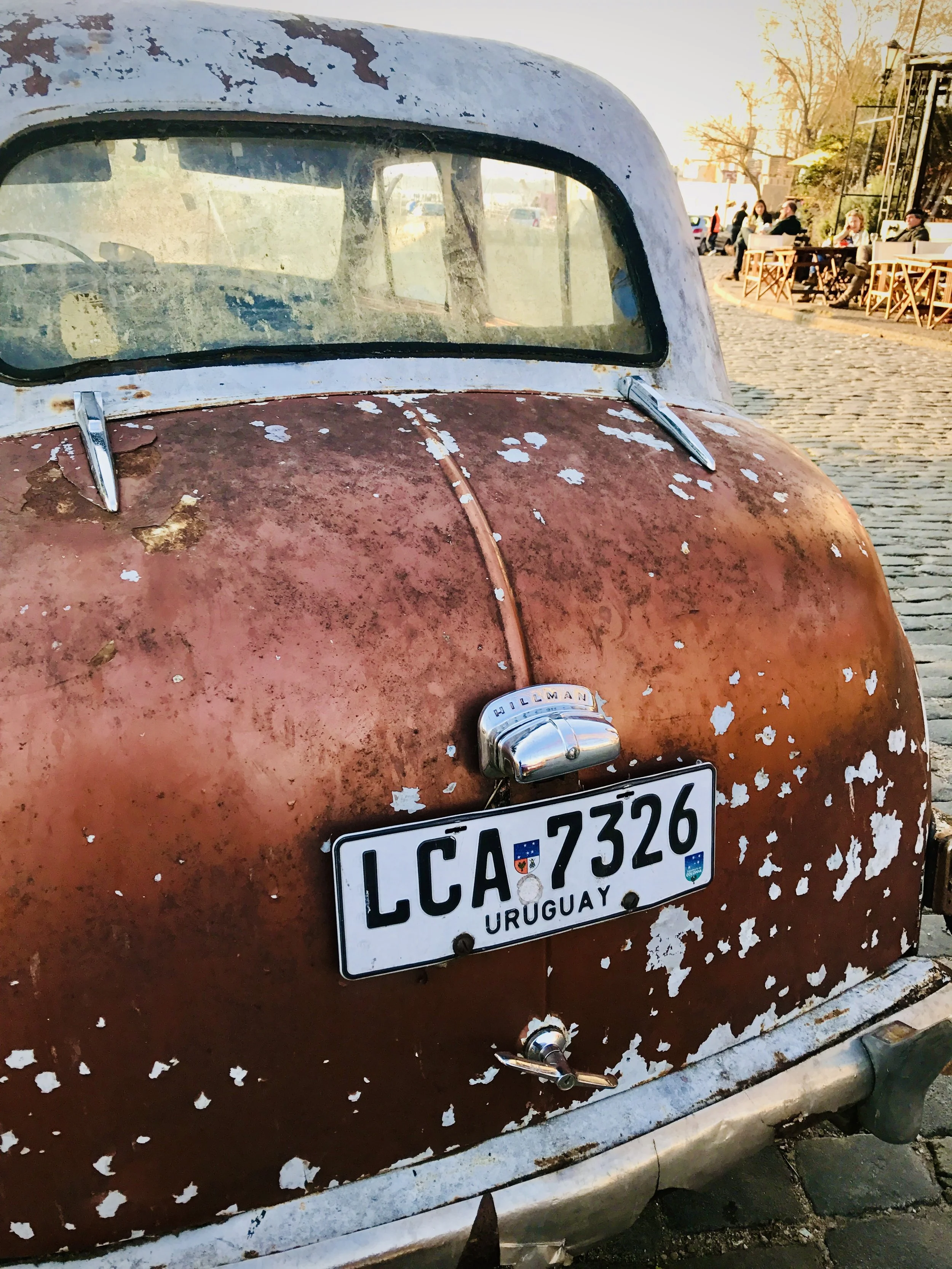 An old, rusty car with a weathered, chipped paint job and a Paraguayan license plate. The background shows an outdoor café with people sitting at tables on a cobblestone street, during what appears to be late afternoon or early evening.
