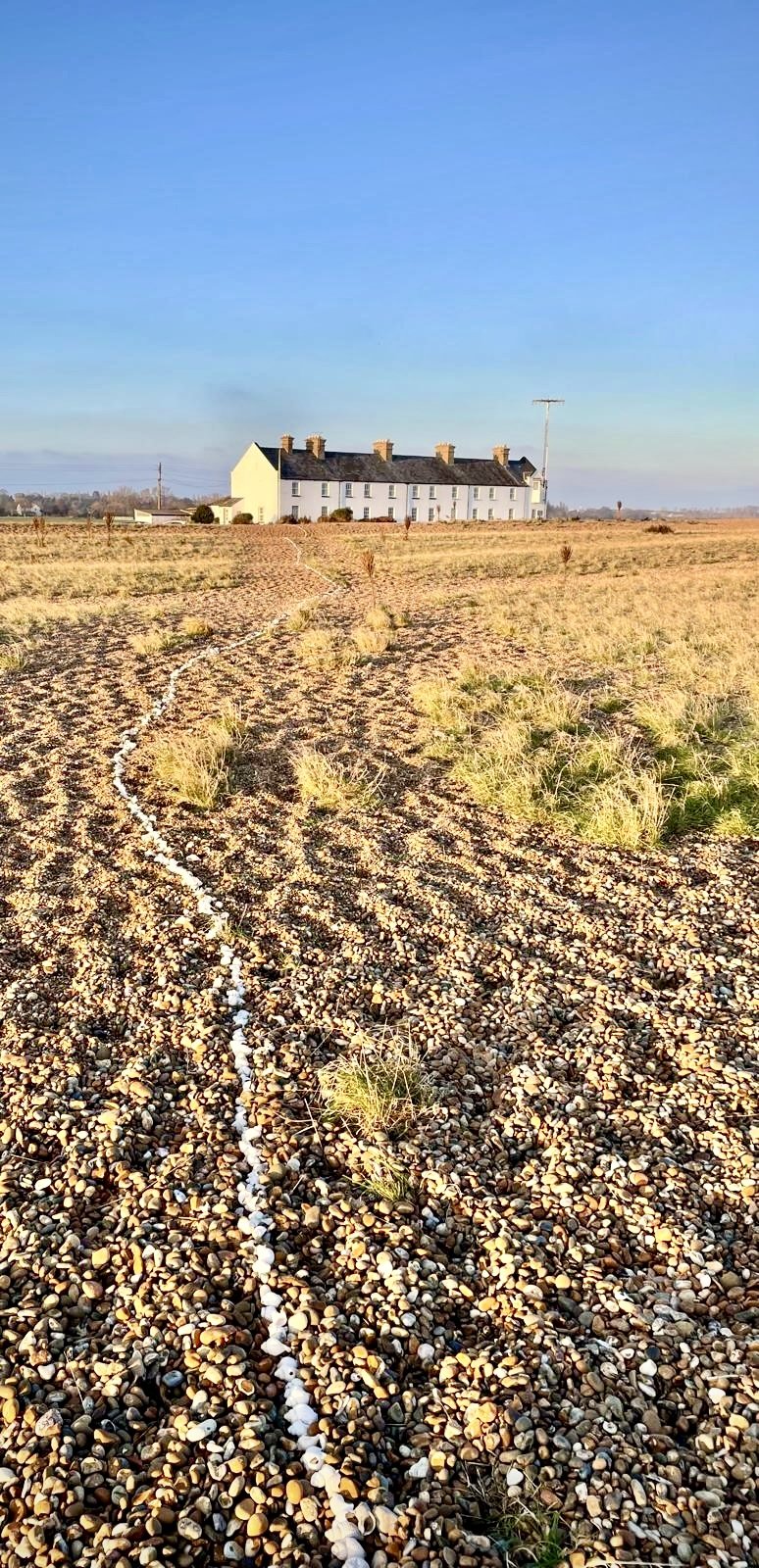 A distant white farmhouse with multiple chimneys in a dry, pebble-covered field under a clear blue sky.