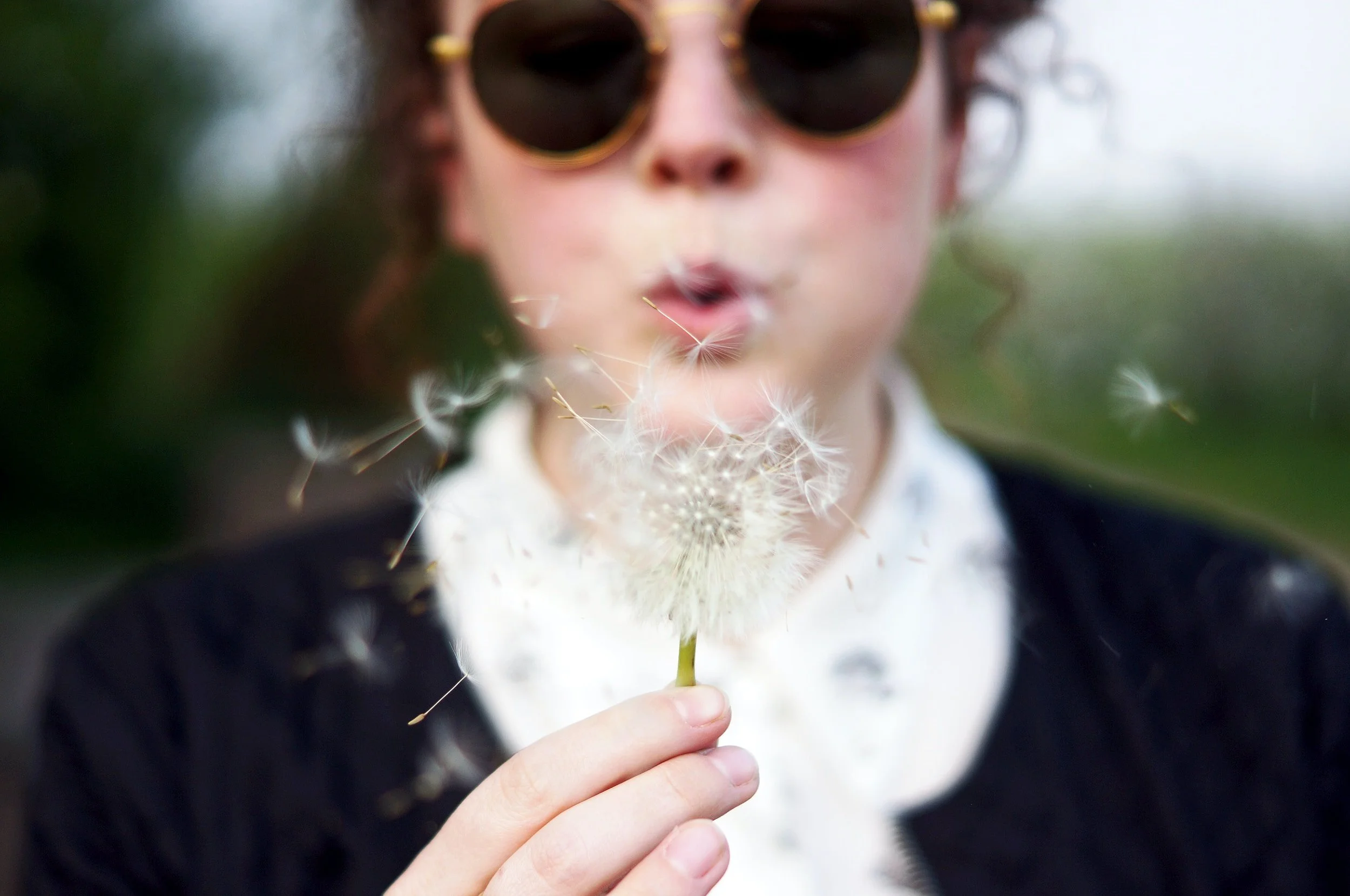 A person wearing sunglasses blowing on a dandelion seed head, with the seeds dispersing into the air.