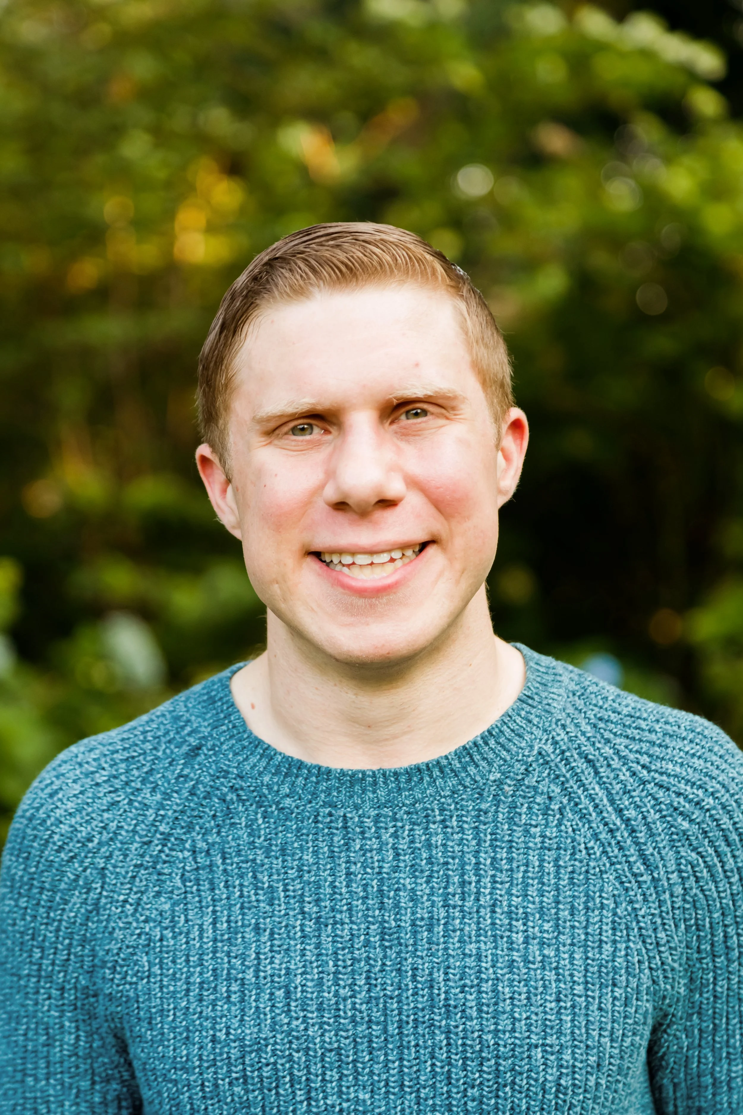 Close-up of smiling young man with short, light brown hair, wearing a blue textured sweater, standing outdoors with blurred green foliage in the background.