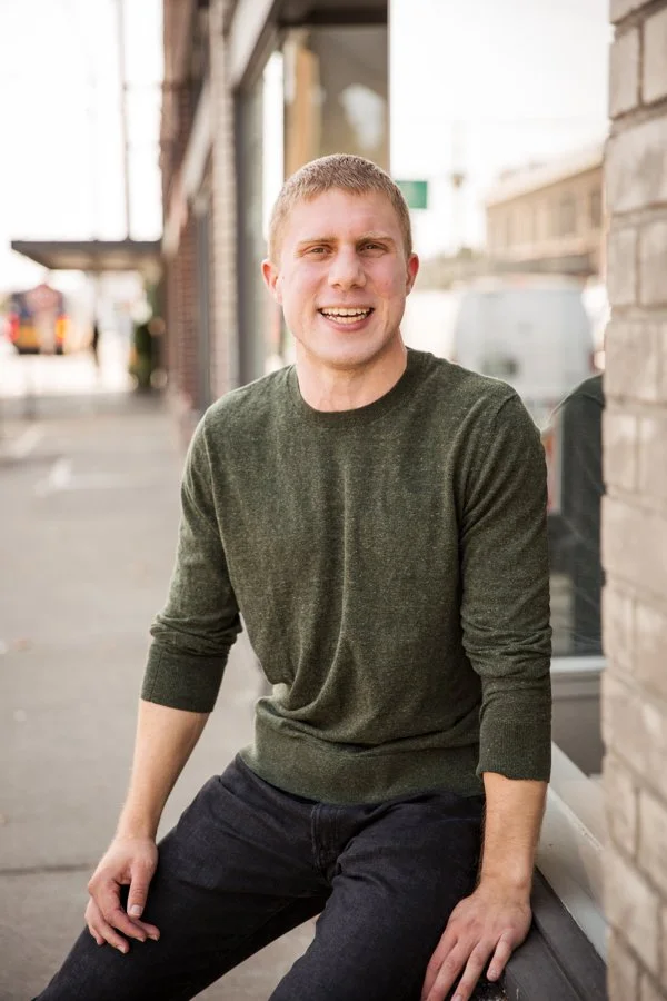 A young man with short blond hair and a friendly smile, wearing a dark green sweater and black pants, sitting on a ledge outside a building in an urban area.