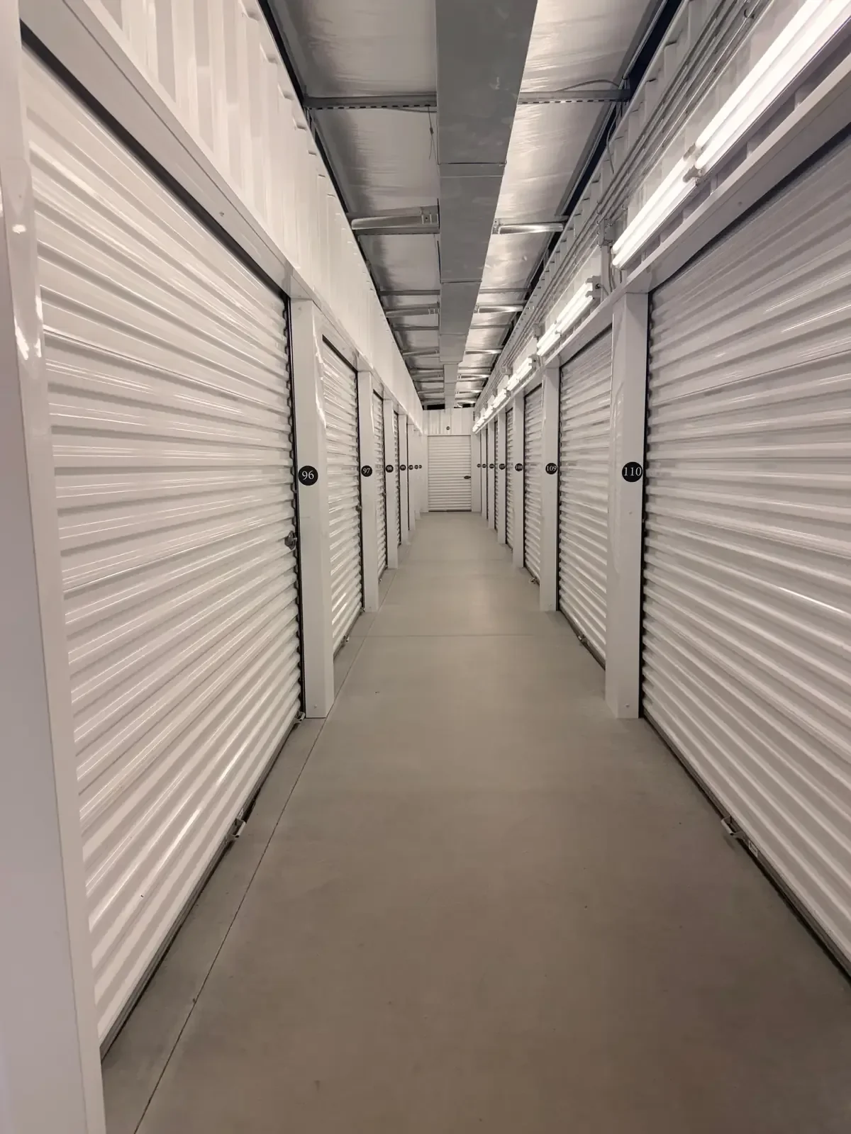 A hallway of storage units with white roll-up doors, each numbered, in a self-storage facility.