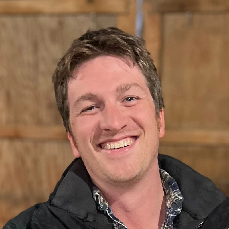 A smiling man with short brown hair, wearing a plaid shirt and a dark jacket, standing indoors with wooden walls in the background.