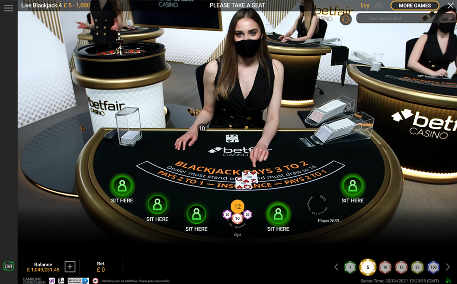 Interior of a casino with a blackjack table showing a dealer with long brown hair and a black face mask. The table has a few players, and the dealer is dealing cards to a player with a hand showing a ten and a three. There are signs and branding for Betfair Casino, with other tables and staff visible in the background.