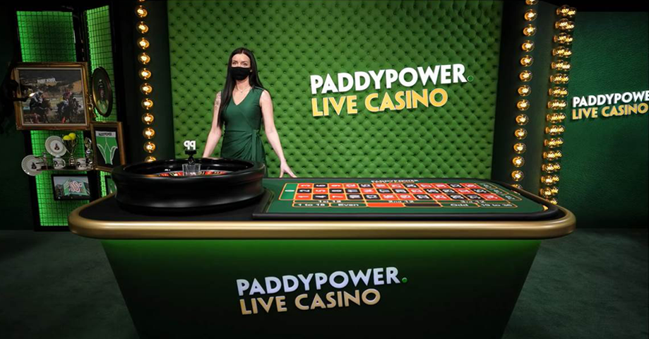 A female dealer standing behind a casino table in a Paddy Power Live Casino studio, with green upholstered walls and a large digital display. She is wearing a black face mask and a green dress, with a roulette wheel and playing cards on the table.