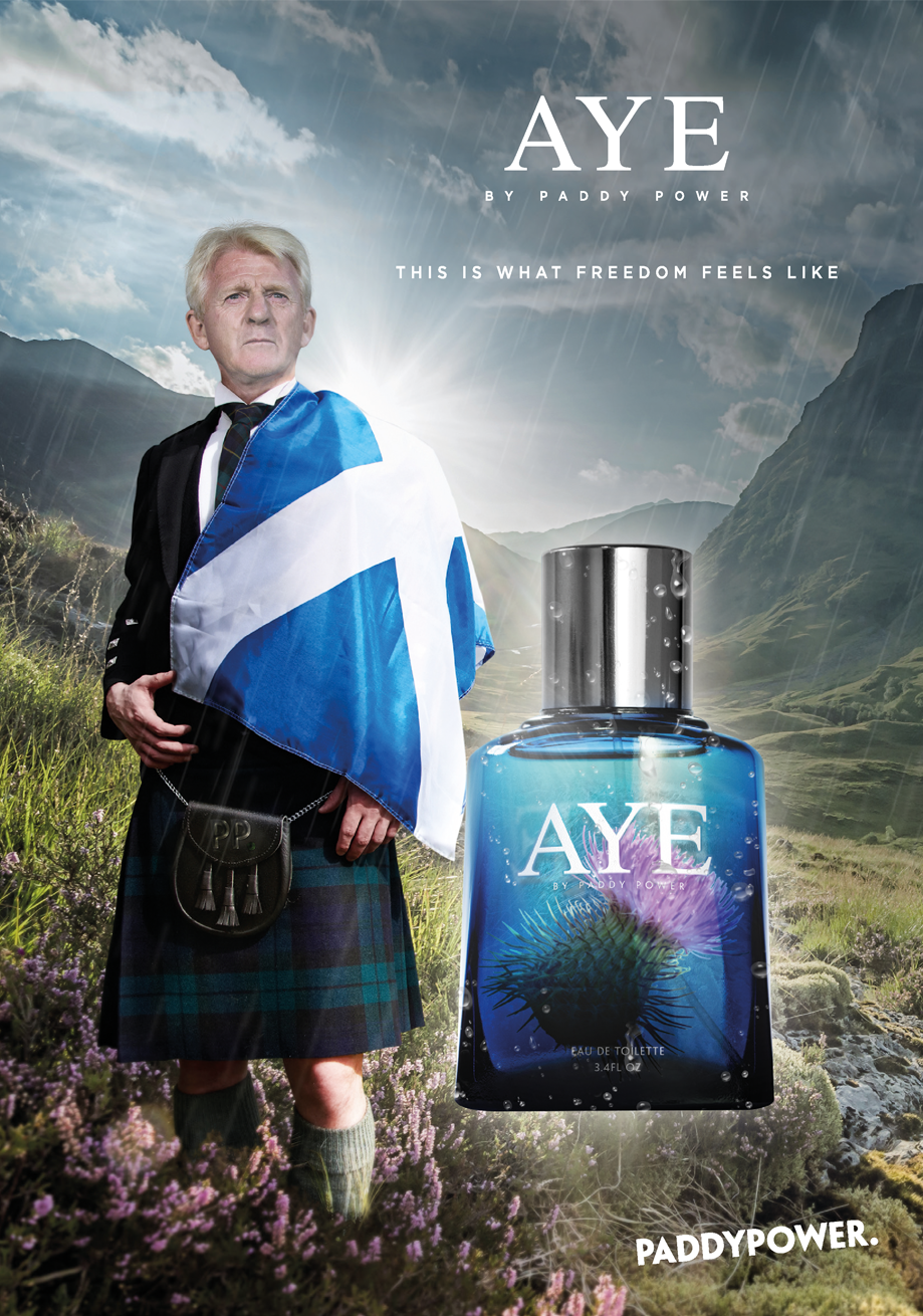A man dressed in traditional Scottish attire, wearing a kilt and a suit jacket, standing in a lush valley with hills in the background. He is holding a Scottish flag draped over his shoulders. A large bottle of AYE by Paddy Power perfume floats next to him, with a landscape of mountains and a cloudy sky behind.