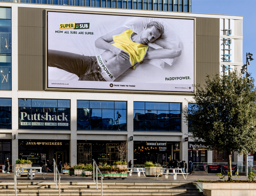 Large outdoor billboard featuring a black-and-white photo of a man lying on his side with a serious expression, wearing a yellow shirt and dark jeans, with the text 'Super Sub' and 'Now all subs are super'.