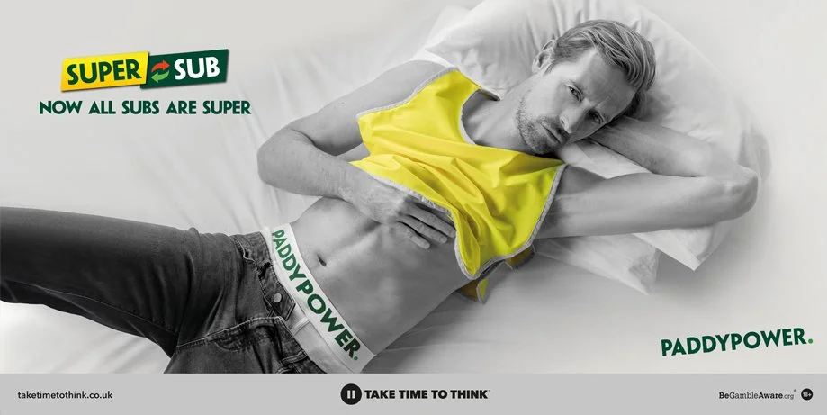 A man lying on a bed, wearing black jeans and a yellow sports vest, with his hand on his stomach. The image features promotional text for Subway and Paddy Power.