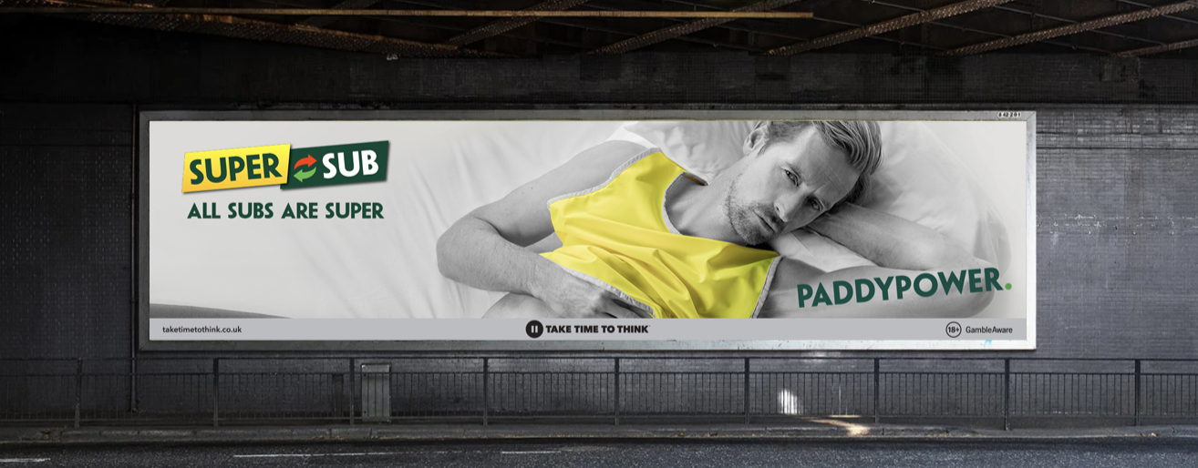An advertisement billboard displaying a man lying in bed, wearing a yellow shirt, with various sports-related phrases and logos, including 'Super Sub,' 'All Subs Are Super,' 'Paddypower,' and a call to action to 'Take Time to Think'.