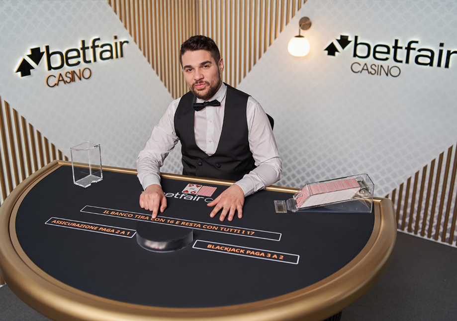 A dealer at a blackjack table in a Betfair Casino setting, wearing a white shirt, black vest, and bow tie, with a deck of cards and poker chip in front of him.