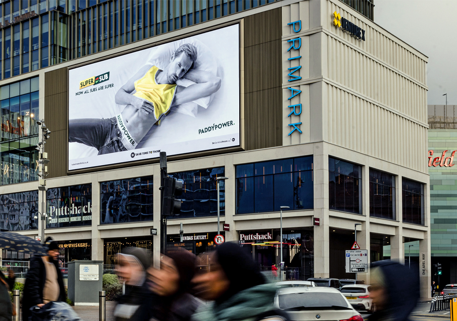 City building with large digital billboard featuring a black-and-white photo of a man lying on a bed, wearing a yellow shirt, with text advertising Super Sub and Paddy Power; the building has signs for Primark, Puttshack, and other shops, with pedestrians crossing the street in front.