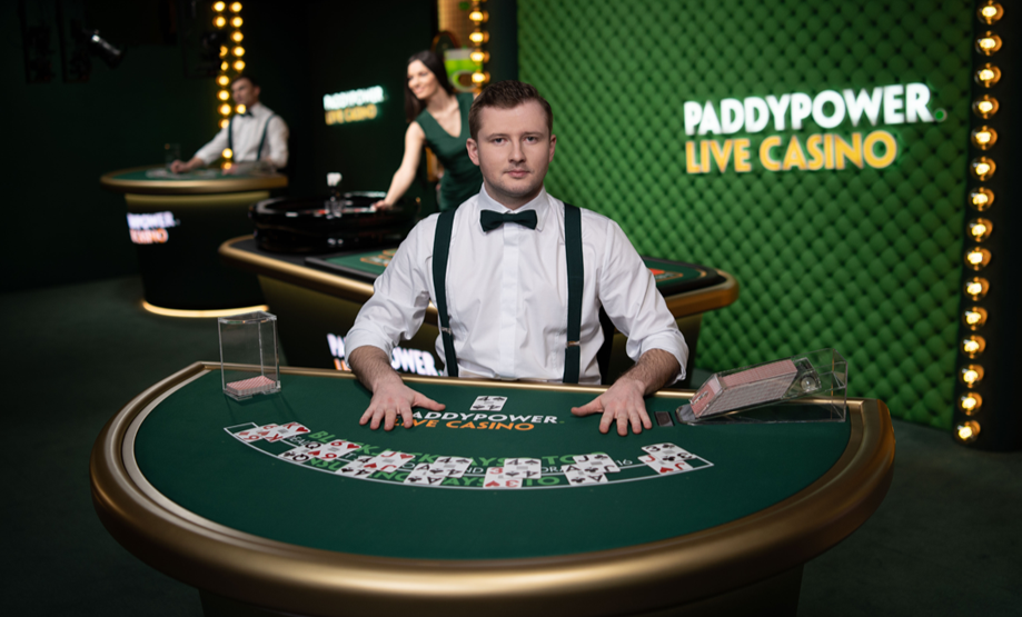 A male casino dealer dressed in a white shirt with suspenders and a bow tie, sitting at a green poker table in a casino with a