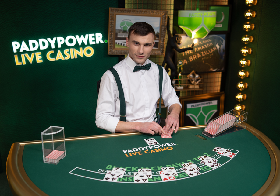 A male dealer at a casino table with playing cards, chips, and a green felt surface, promoting Paddy Power Live Casino.