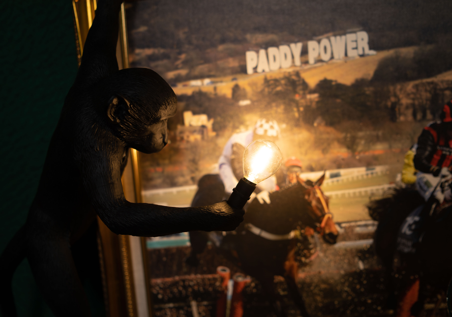 A figurine of a black cat holding a light bulb, positioned in front of a horse race poster with the sign 'Paddy Power' in the background.