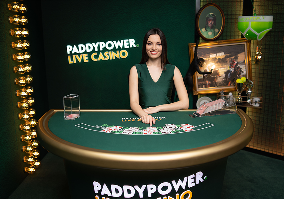 A woman standing behind a blackjack table at Paddy Power Live Casino, smiling at the camera. The background features the Paddy Power Live Casino logo, a framed picture, a large green cup, and a small black monkey figurine.