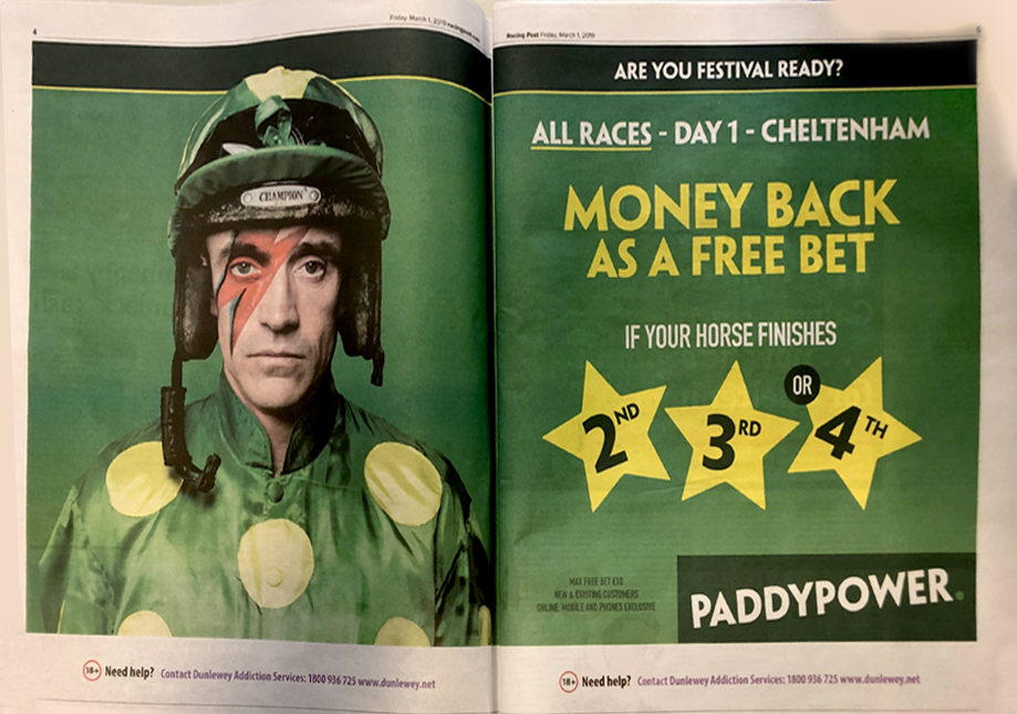 An open magazine page featuring a man dressed as a jockey with painted lightning bolt makeup on his face. The left side shows him in a green jockey outfit and helmet. The right side contains promotional text about a racing festival in Cheltenham, offering money back as a free bet if a horse finishes in second, third, or fourth place.