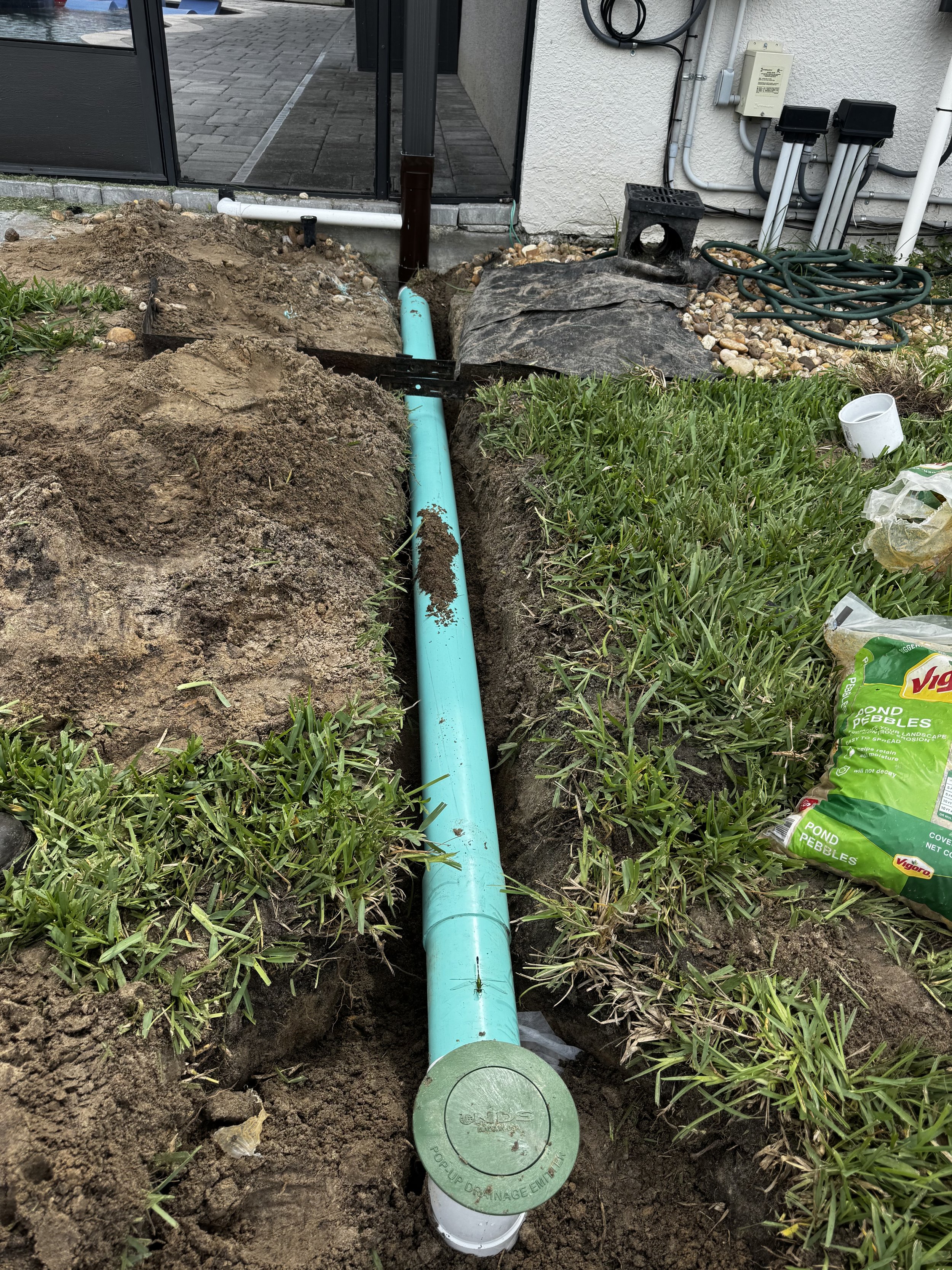 A blue underground sewer or storm drain pipe installed in a trench in a yard with grass on either side. The pipe is connected to a round cap labeled 'PUMP UP DRAINAGE EMITTER.' Nearby, there is a black plastic drainage cover, a black crate, a coiled hose, and some gardening supplies, with a house wall and electrical wiring in the background.