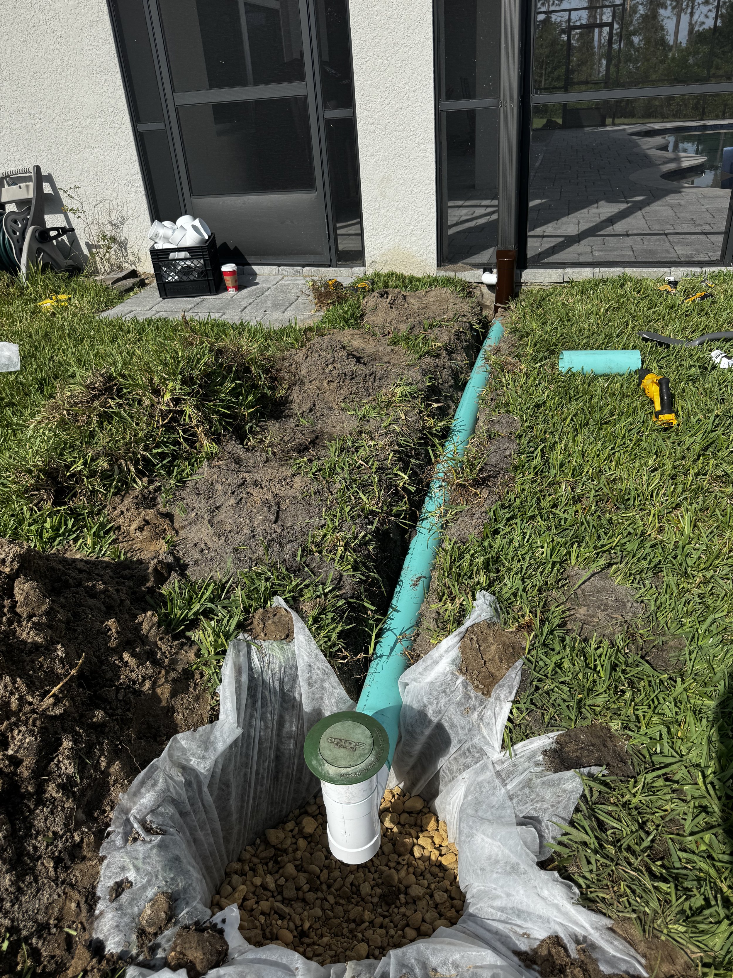 A plumbing installation in progress outside a house, showing a trench with gravel, a white pipe, and a teal pipe connected to a brown fitting, surrounded by green grass, with tools and supplies nearby.