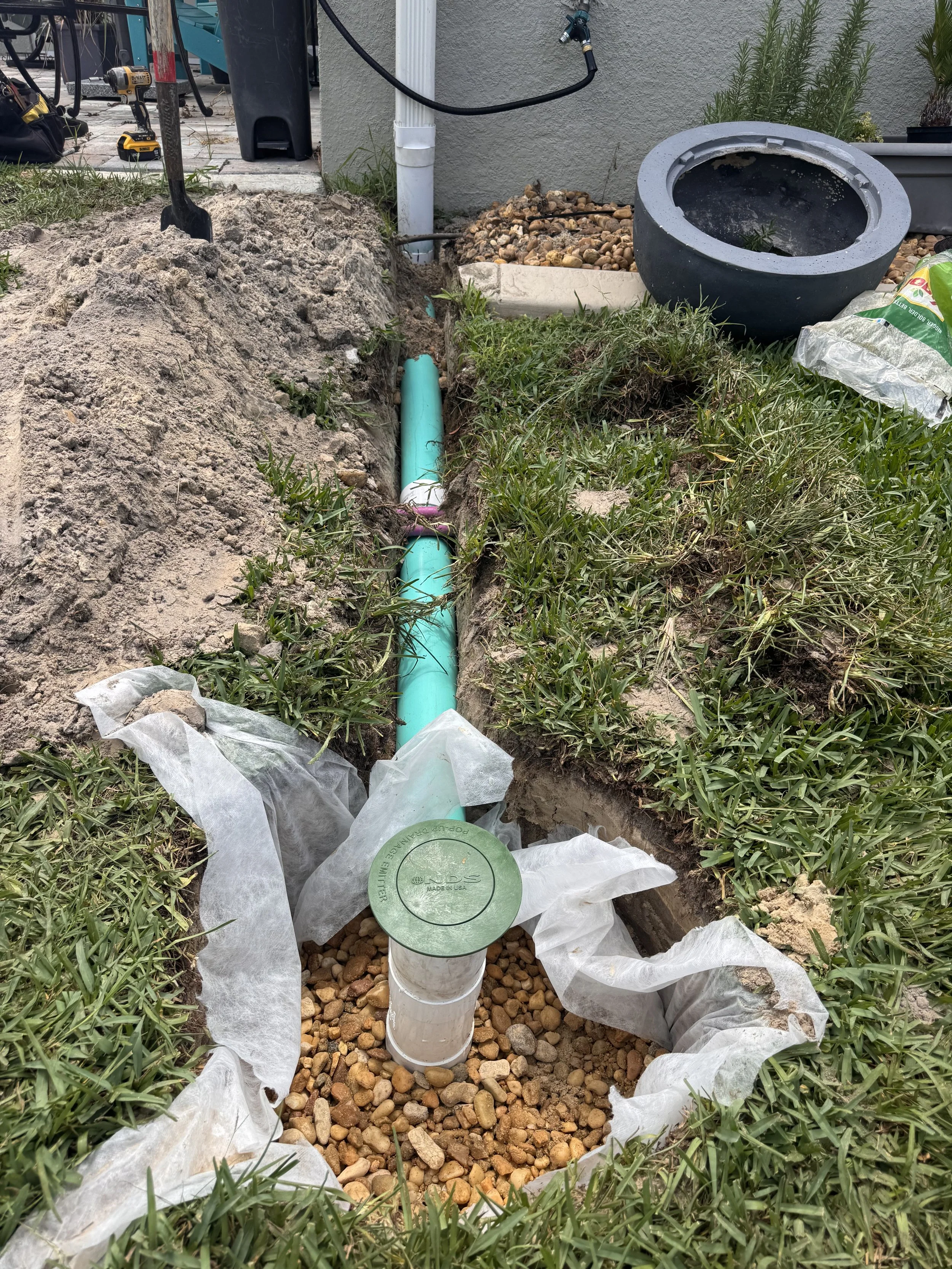 A garden or yard with a trench dug for underground pipe installation. A blue pipe runs vertically through the trench, which is lined with white fabric for weed barrier. Gravel covers the bottom of the trench. Nearby are tools, a black planter, and various gardening supplies.