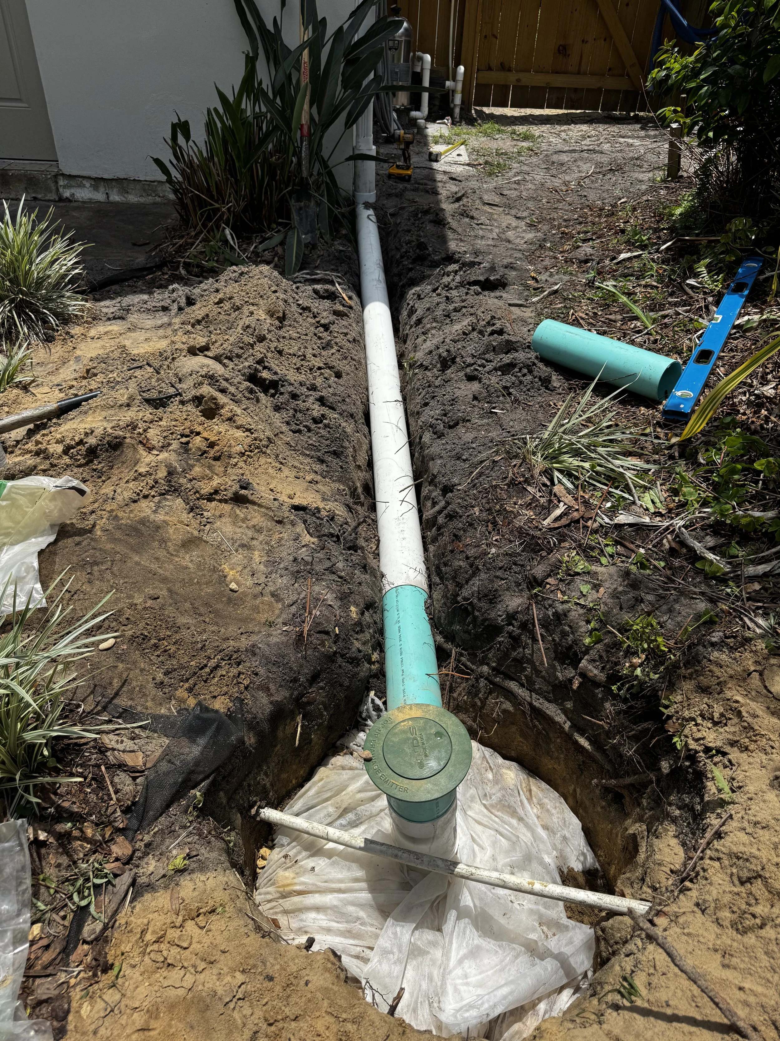 Underground plumbing installation with pipes and tools in a trench outside a house, with a wooden fence in the background.