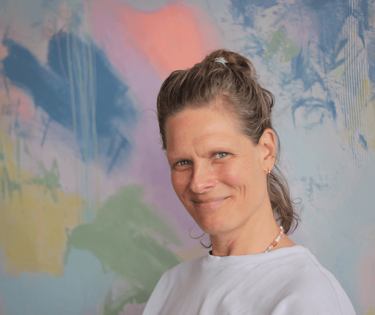 Eine Frau mit braunen, geflochtenen Haaren, die einen weißen T-Shirt trägt, vor einer bunten, abstrakten Wandgestaltung.
