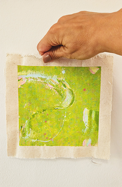 Hand hält ein Stück Stoff mit abstraktem, grünen, pinken und weißen Muster gegen eine weiße Wand.