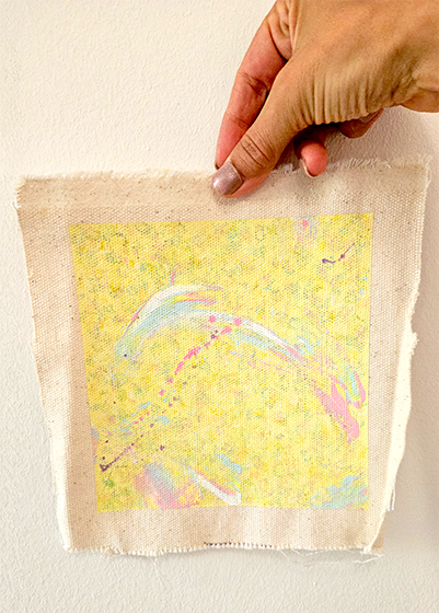 Eine Hand hält ein Stück Stoff mit einem bunten, abstrakten Muster in Pastellfarben, das auf einem beige Hintergrund gedruckt ist.