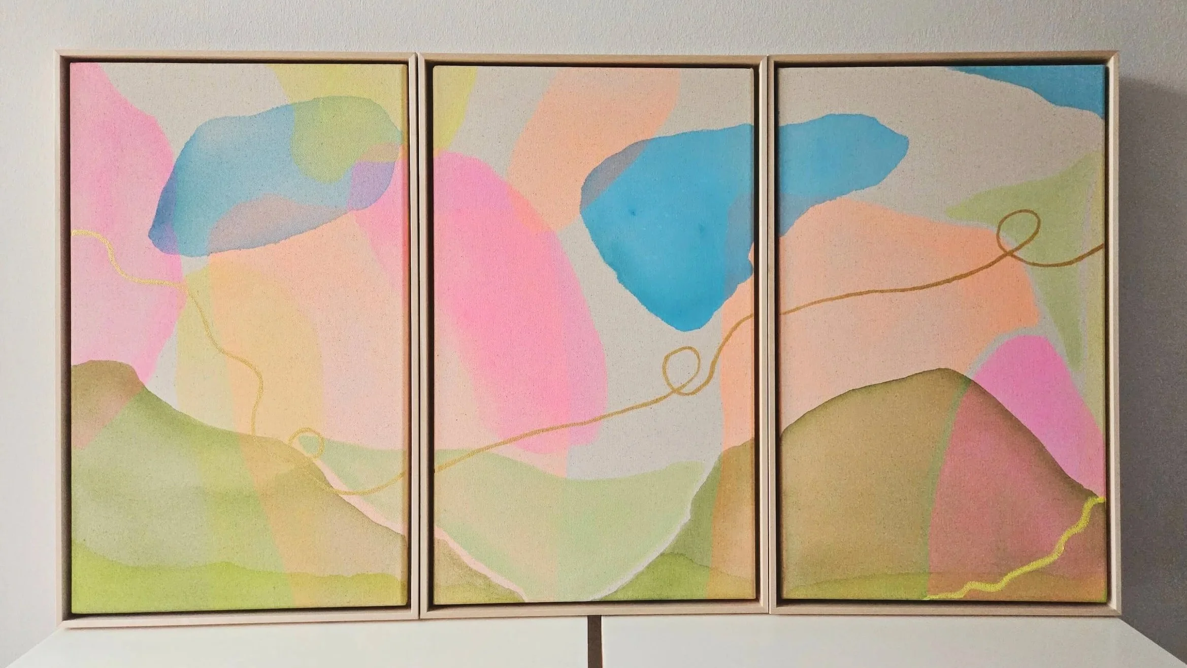Drei nebeneinander stehende rechteckige Bilderrahmen mit abstrakten Pastellfarbenen Gemälden, hauptsächlich pink, blau, grün und beige, mit fließenden Linien auf einer weißen Wand befestigt.