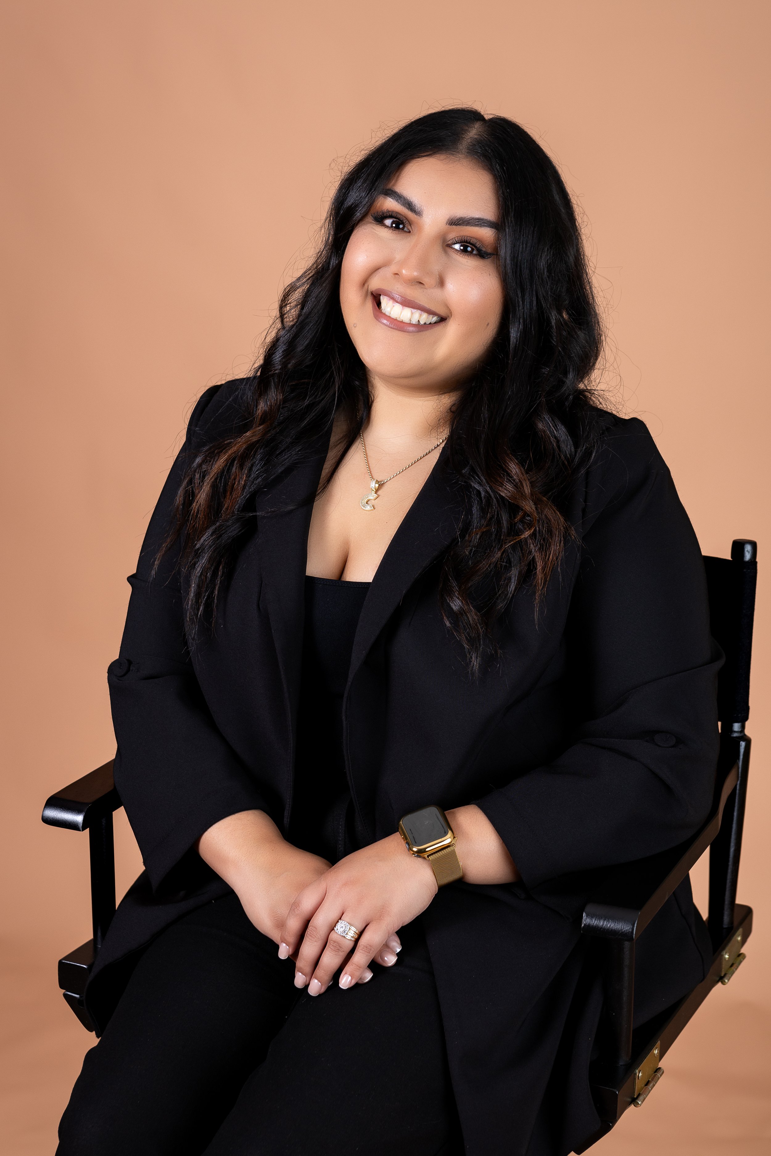A woman with long black hair, wearing a black blazer and shirt, sitting on a black chair against a beige background. She is smiling, showing her teeth, and has a gold necklace with a 'C' pendant, a gold watch, and a ring on her left hand.