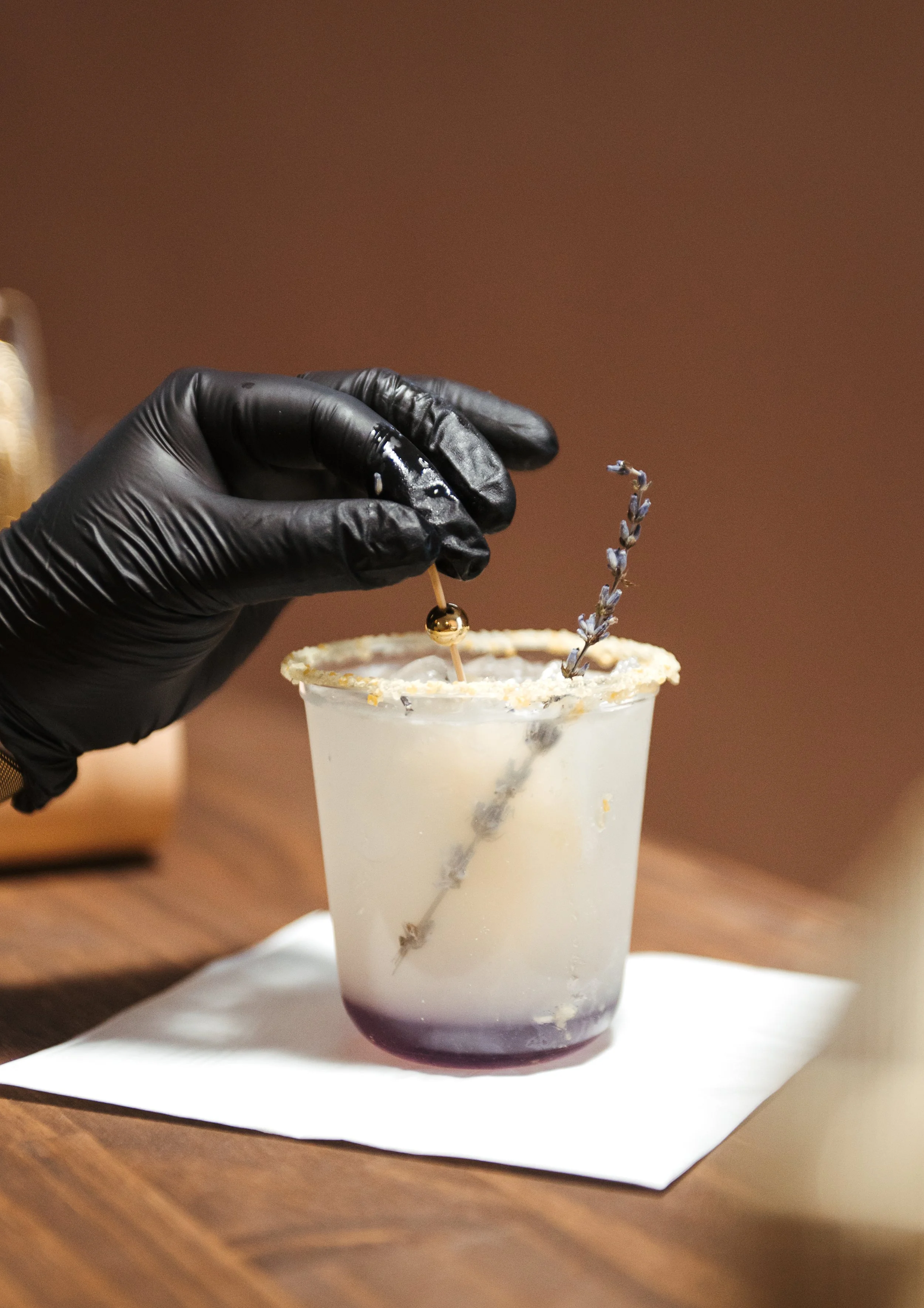 A hand wearing a black glove holding a cocktail stick with a small gold ball on top, inside a glass decorated with lavender and sugar around the rim, on a wooden bar top with a white napkin underneath.