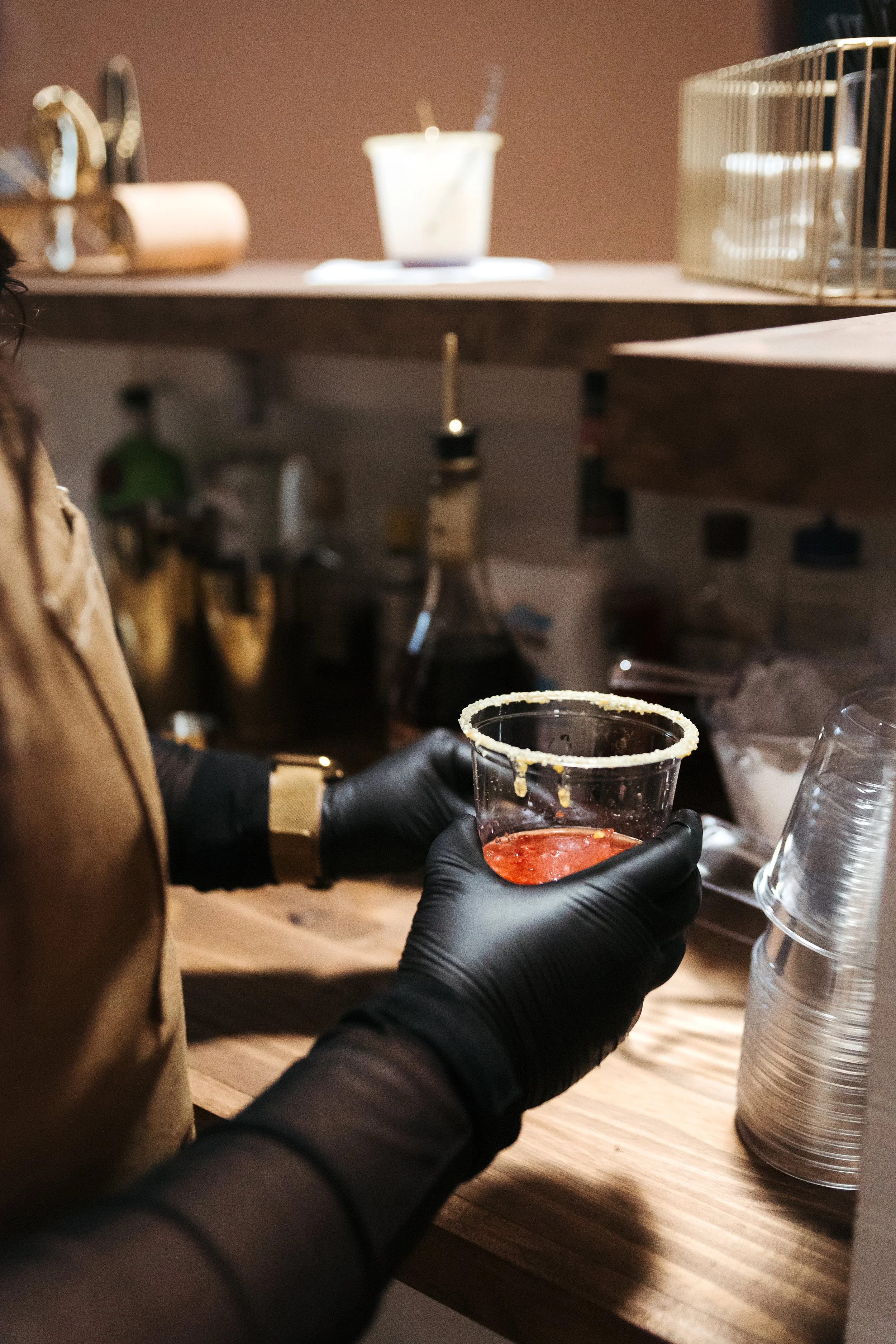 A person wearing black gloves holding a plastic cup with a salted rim and red liquid inside, possibly a cocktail, in a busy kitchen or bar setting with various bottles and stacks of clear plastic cups.