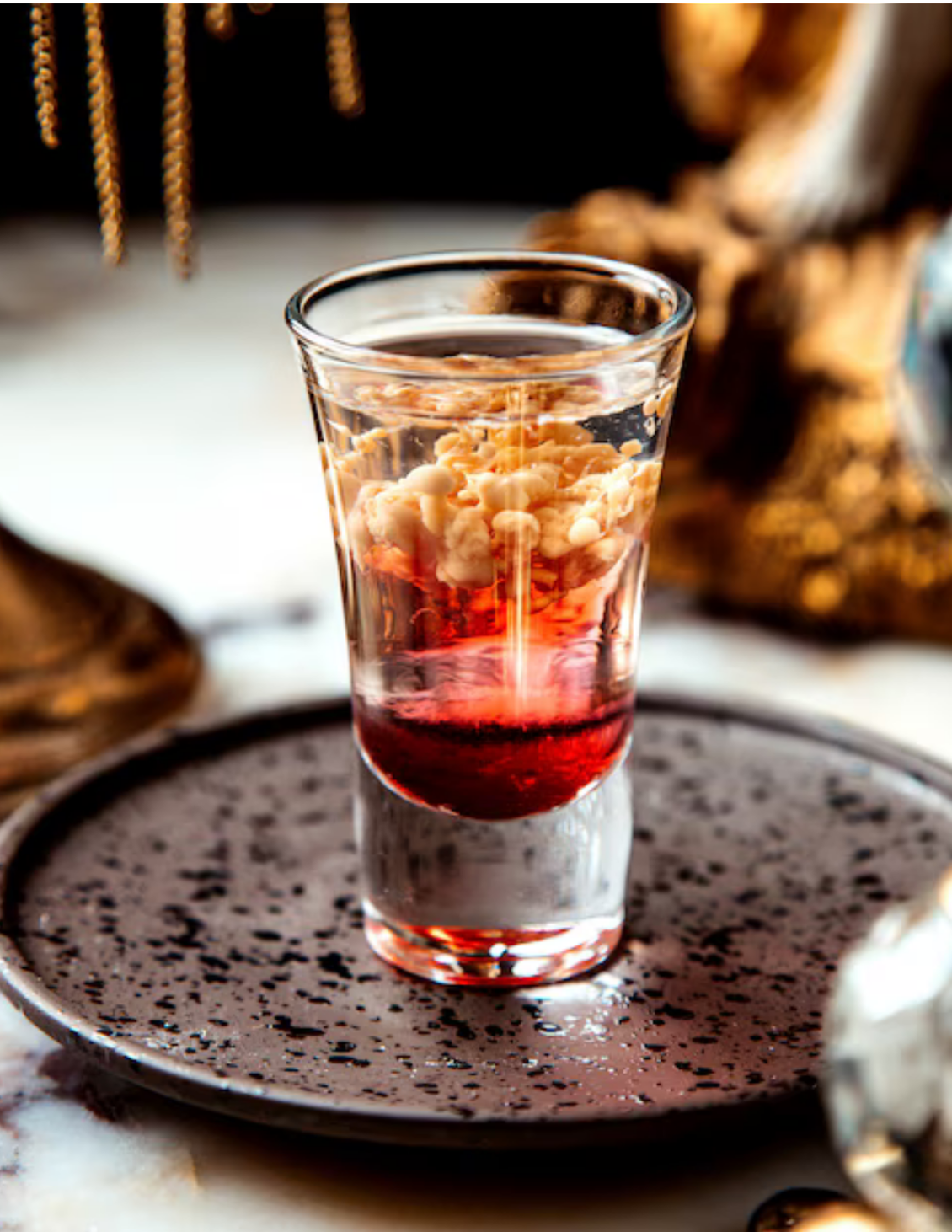 Shot glass with a layered cocktail containing red and white ingredients, placed on a speckled plate.