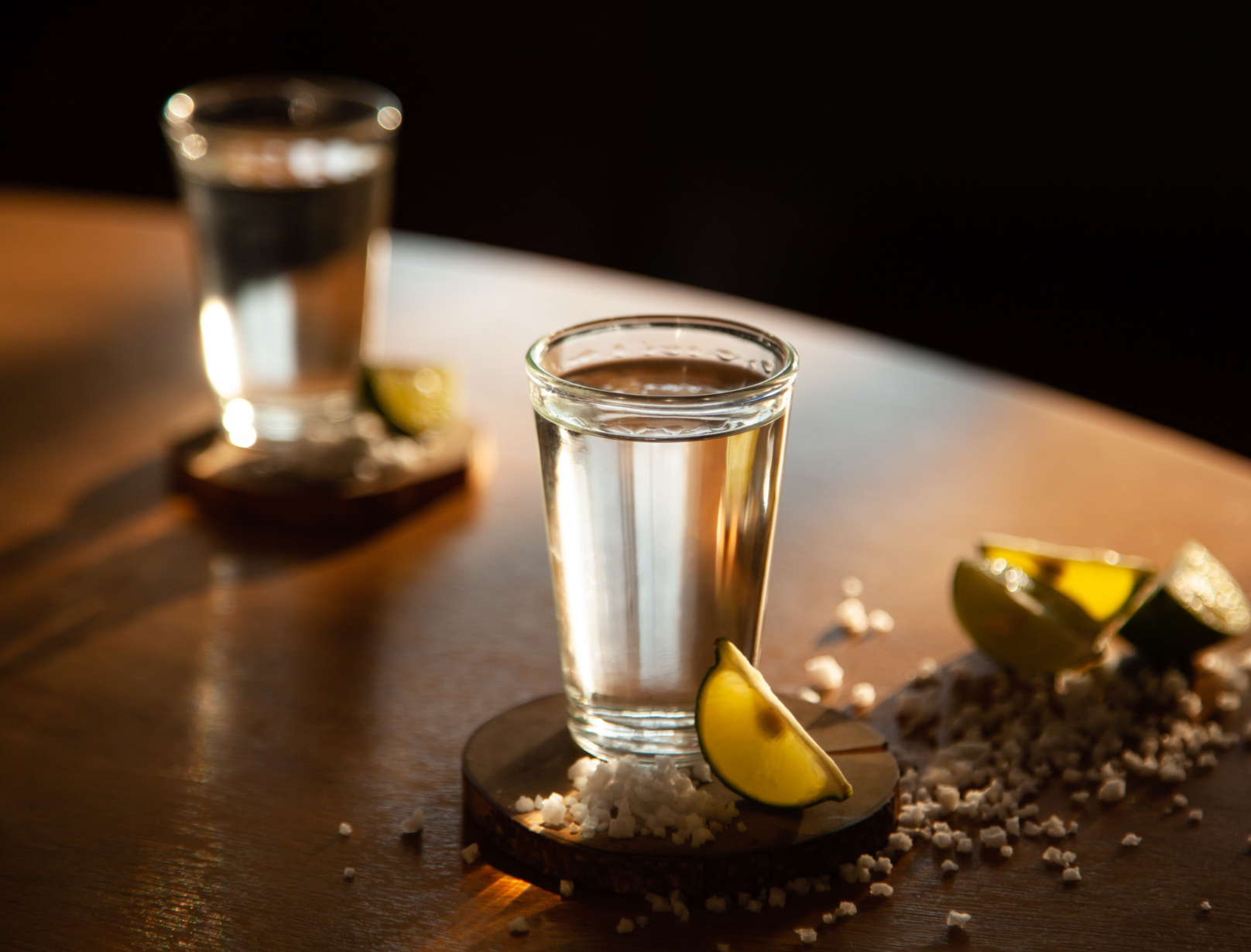 A glass of water on a wooden surface with lemon wedges, salt, and scattered pepper in the foreground, and another glass of water in the background with warm lighting.