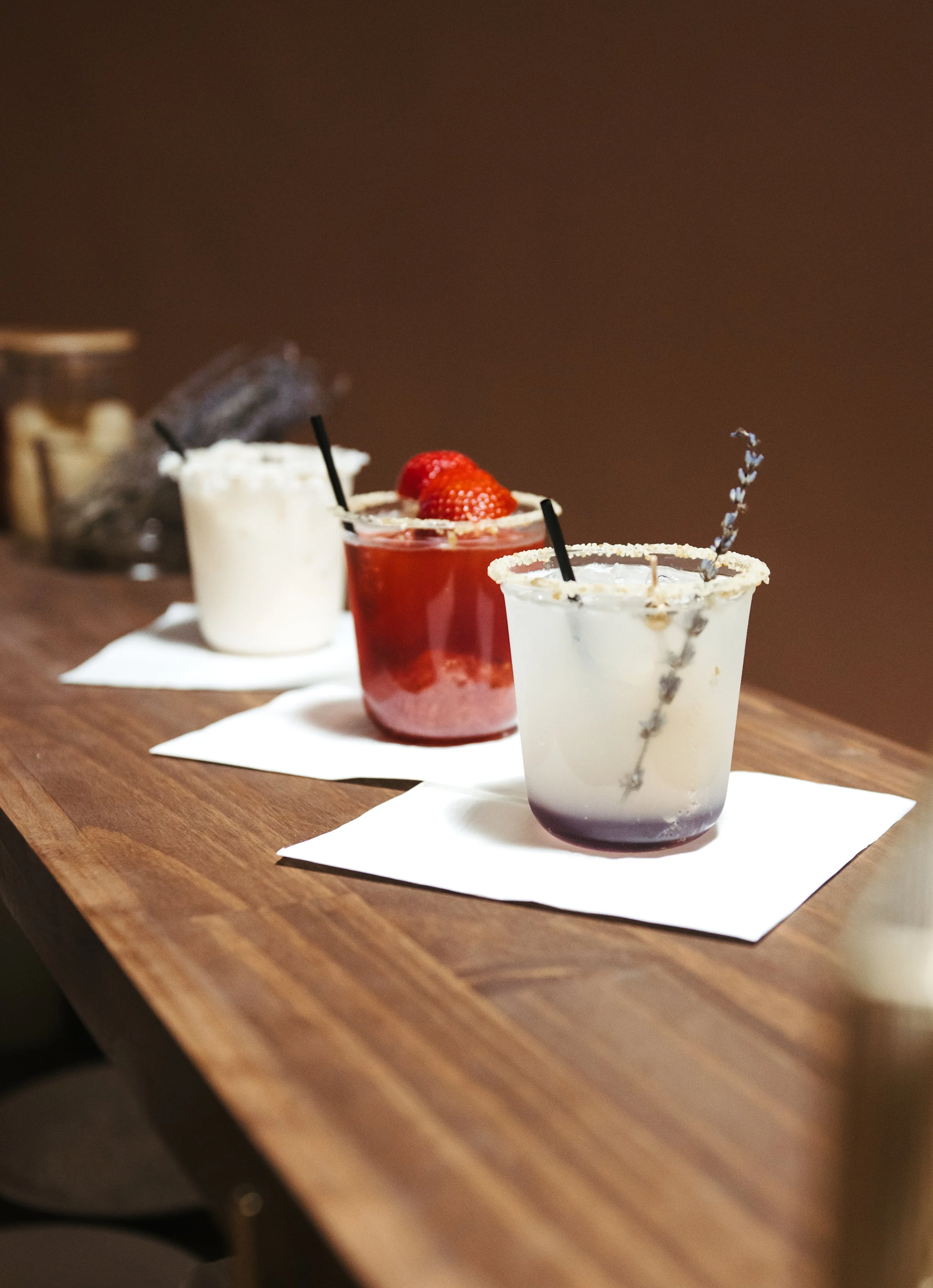 Three cocktails on a wooden bar top, two with white sugar rims and one with a strawberry garnish, all on white napkins.