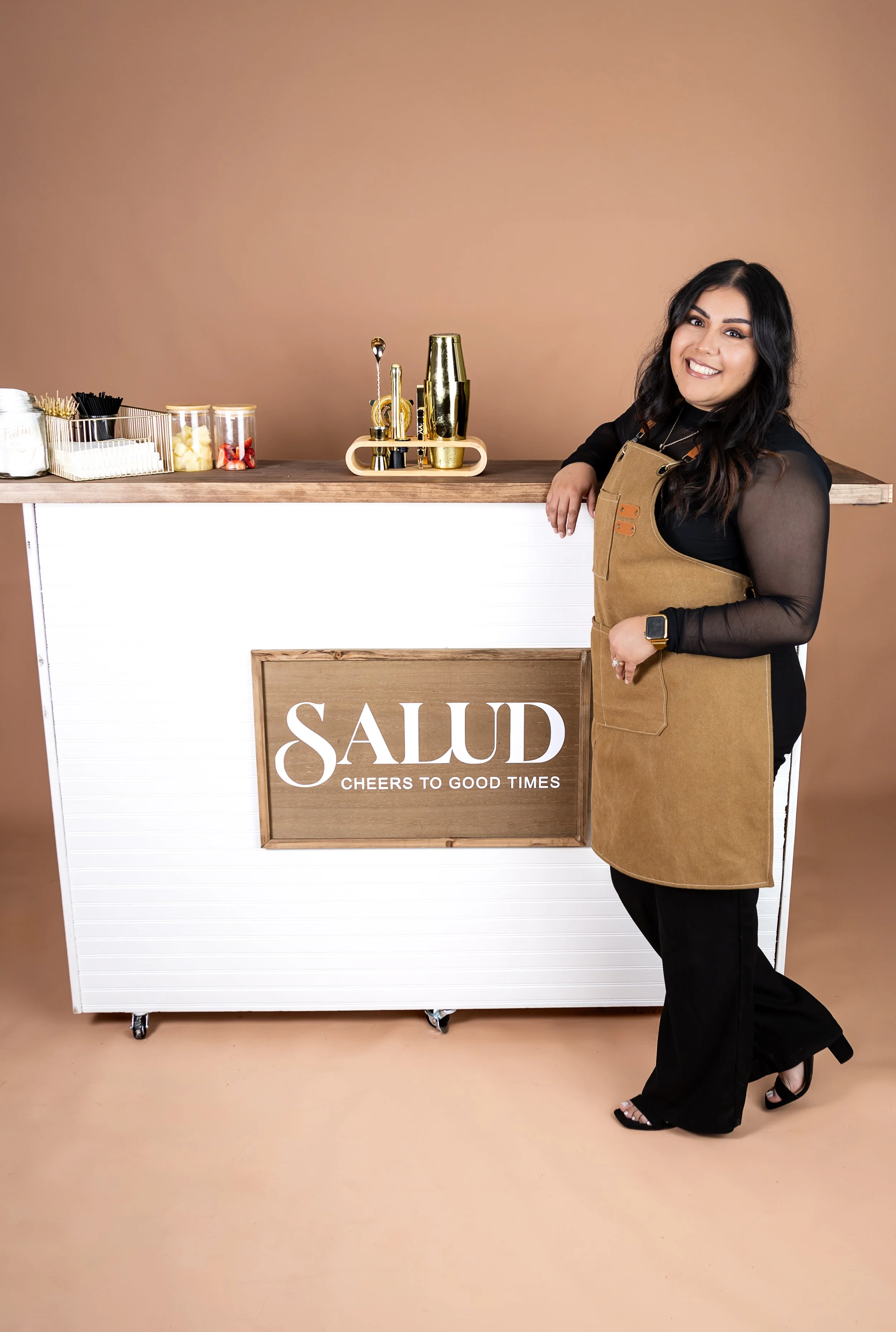 A woman with black hair, wearing a black top, black pants, a tan apron, and high heels, smiling and leaning on a white and wood bar counter with a sign that says 'SAlUD Cheers to Good Times.' In the background, there are various jars and bar tools on a wooden surface against a beige wall.